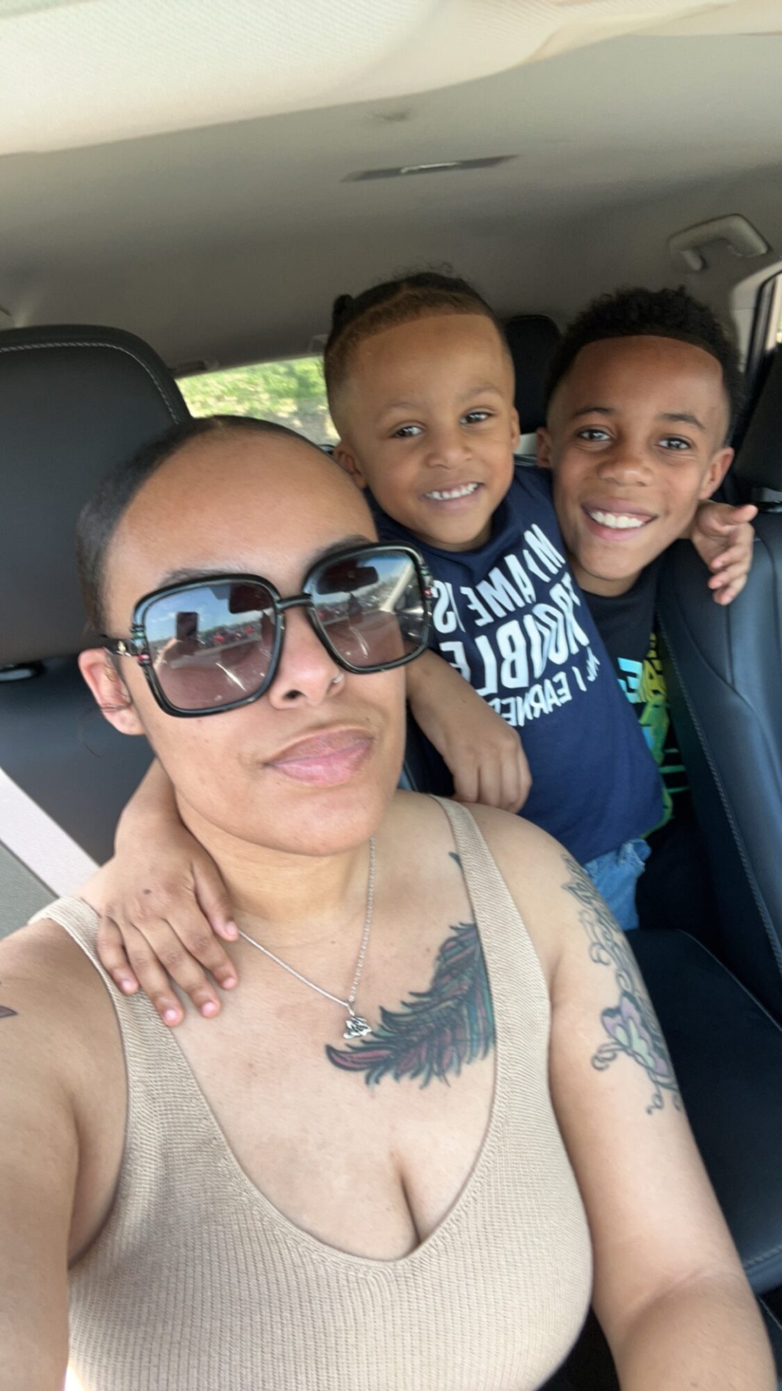 Woman with sunglasses and tattoos in car with two smiling children behind her.