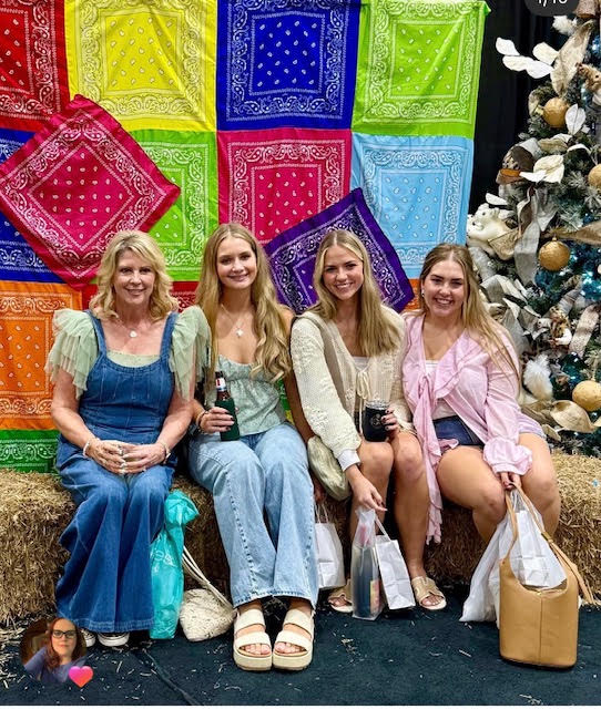 Four women sitting in front of colorful bandanas and a decorated Christmas tree, smiling and holding shopping bags.