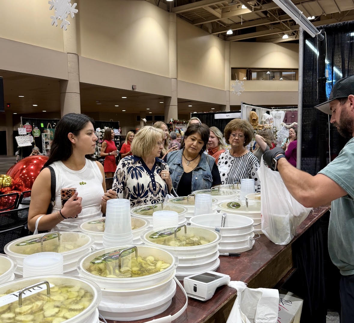 Group of women at a food sampling booth with bowls of pickles, cups, and a man serving food, in a large indoor space.