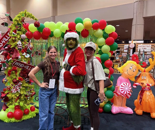 Two women and a person in a Christmas tree costume pose in front of a balloon arch and holiday decorations.