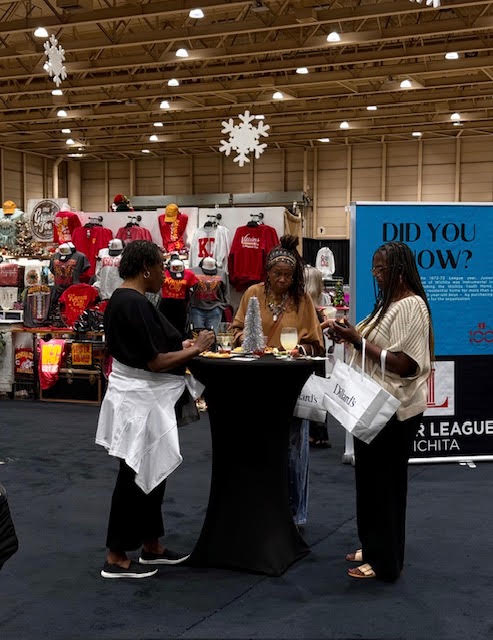 Three women stand around a tall table in a store with holiday decorations and clothing displays in the background.