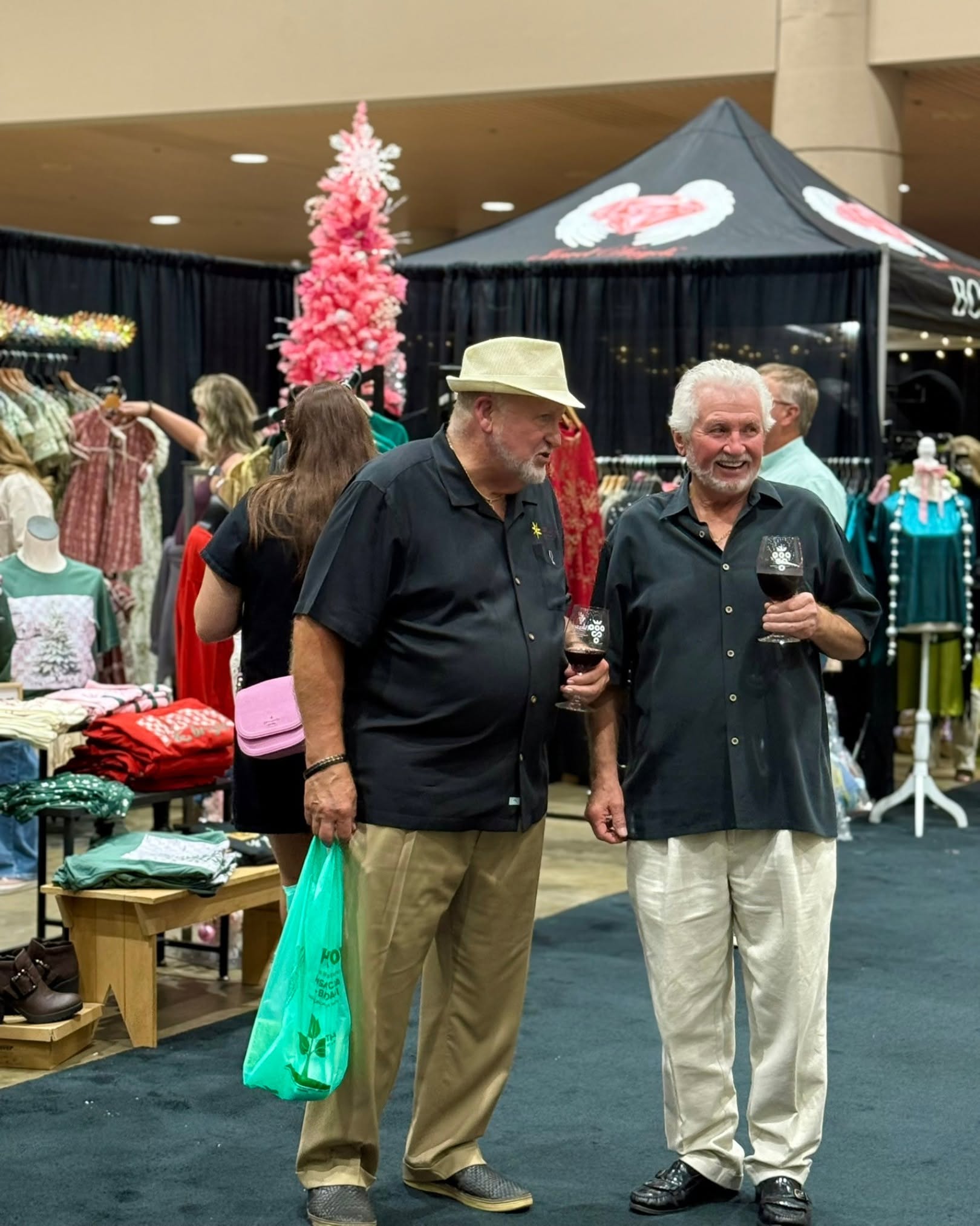 Two men stand and chat at an indoor event, holding glasses of wine, with clothing displays and a pink Christmas tree in the background.