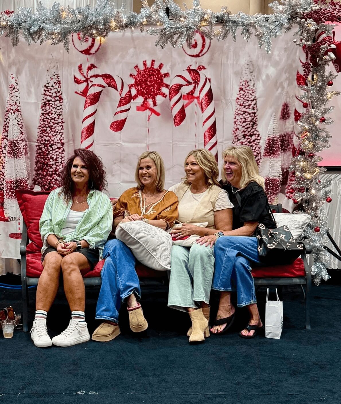 Four women sitting on a red bench in front of a Christmas-themed backdrop with candy canes and snowflakes, smiling.