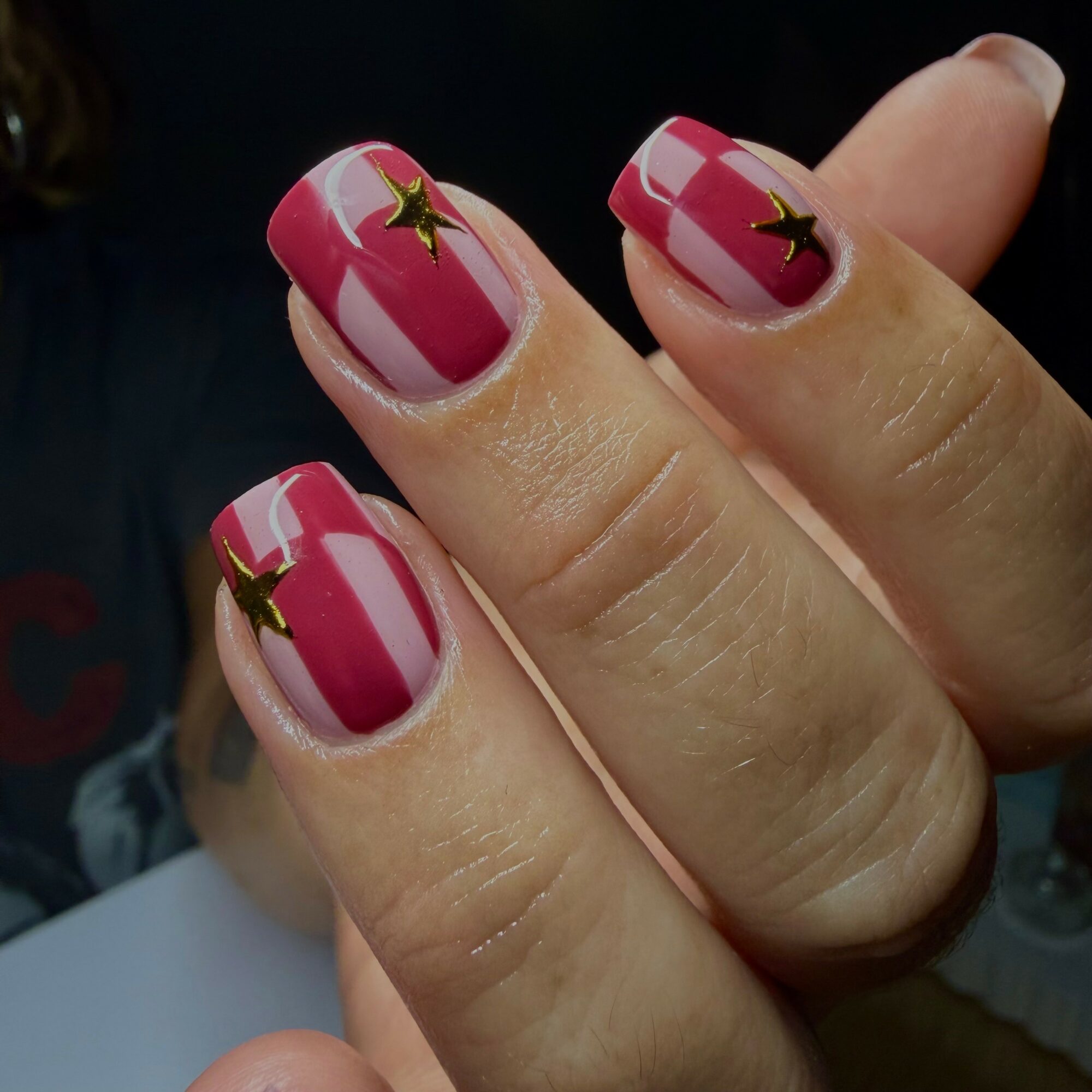 Close-up of fingers with pink and white striped nail polish and gold star decals, against a dark background.