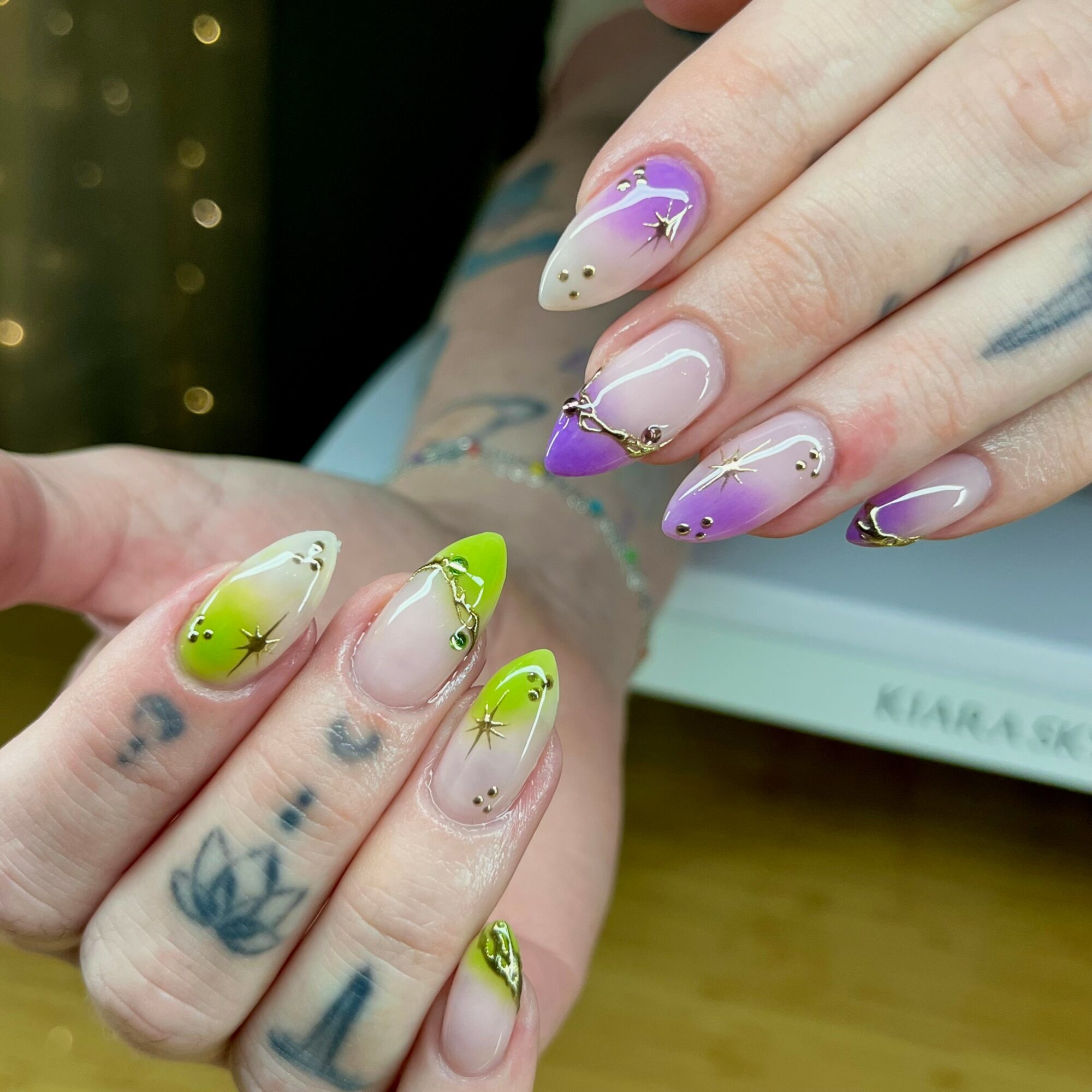 Close-up of hands with colorful, decorated nails and tattoos, showing nail art with purple, green, and beige colors, and small embellishments.