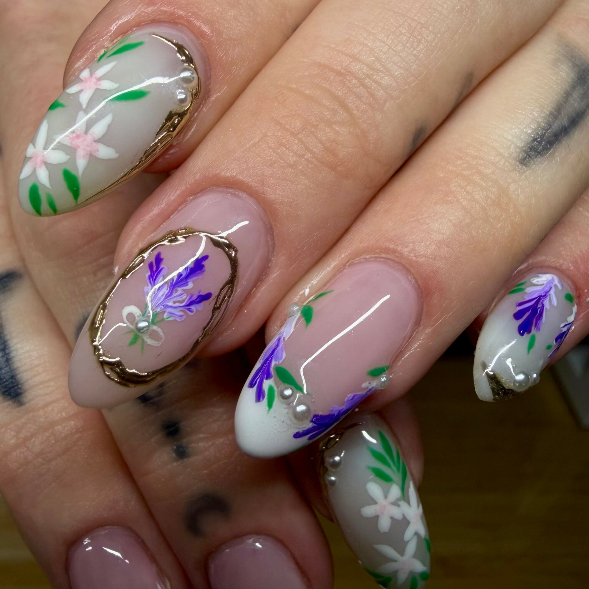 Nail art with floral designs in white, purple, and green on a light pink base, with gold accents.