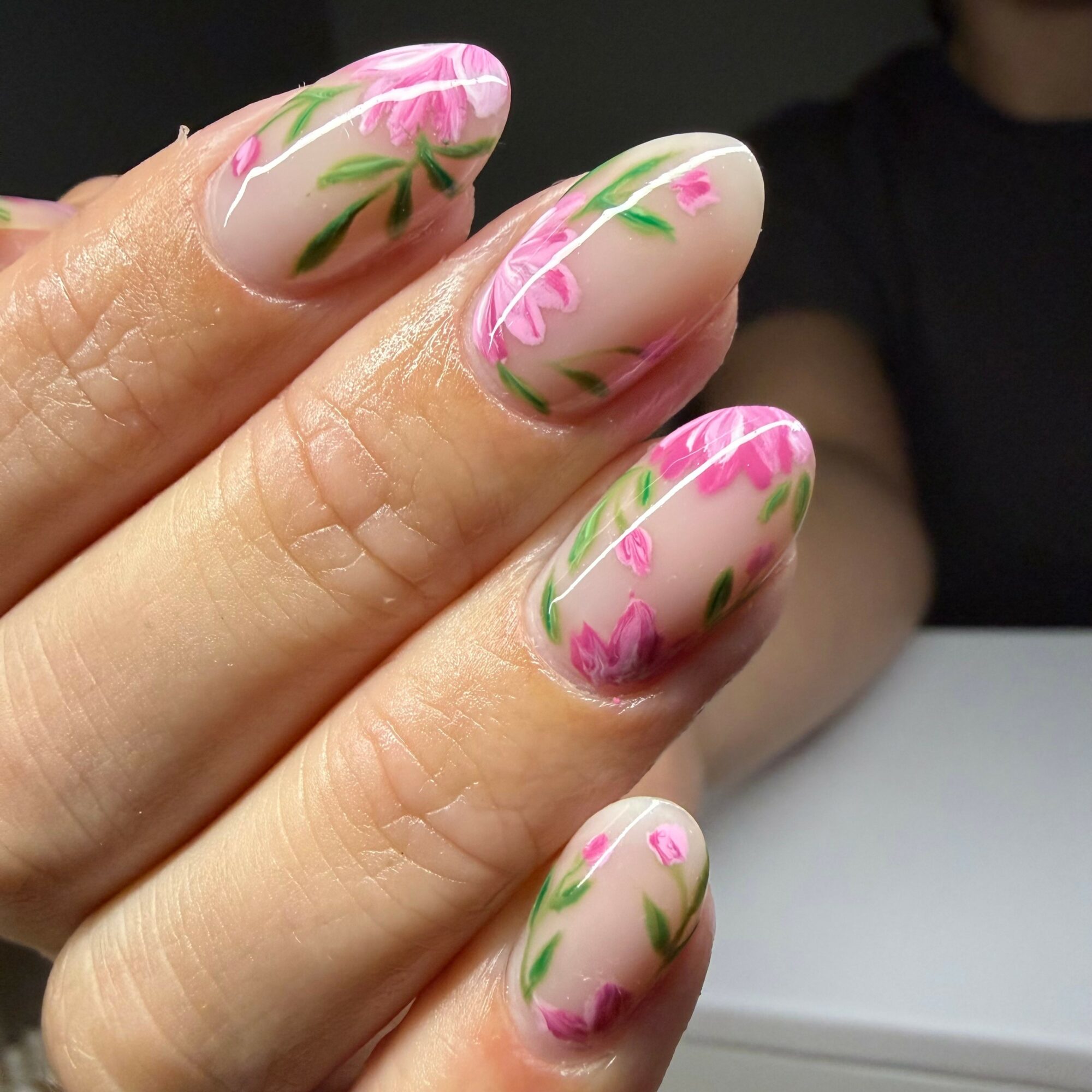Close-up of hand with nails painted with pink flowers and green leaves on a beige background.