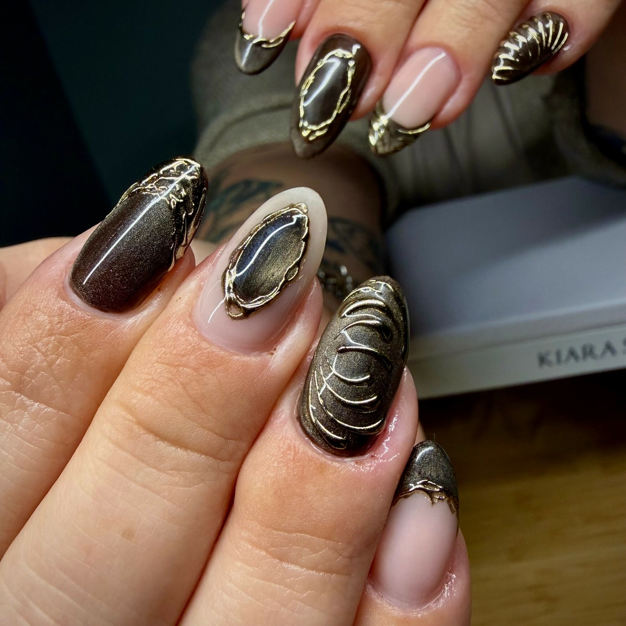 Close-up of hands with decorated nails featuring metallic, black, and beige designs, some with gold accents, on a wooden surface.