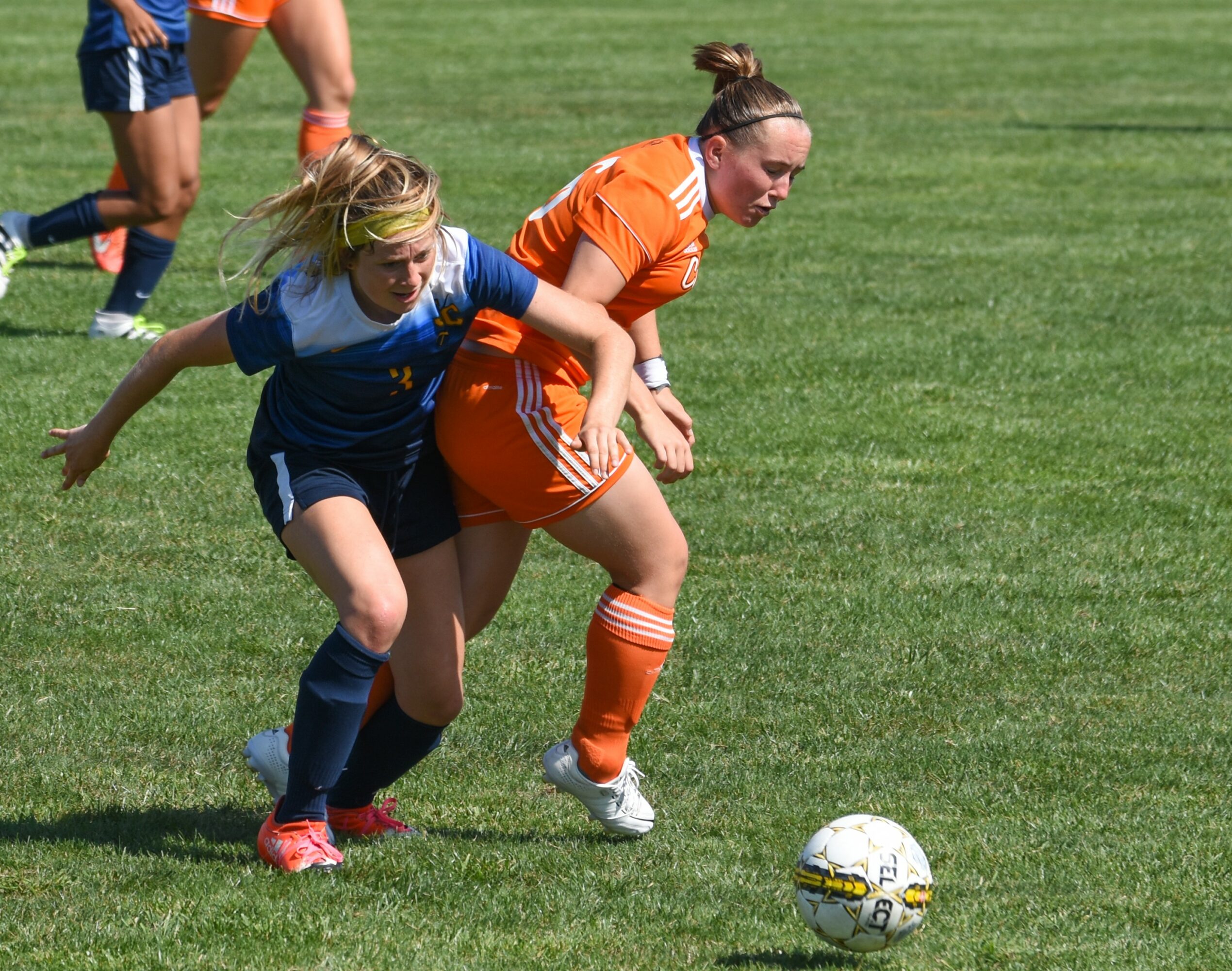 Two female soccer players compete for the ball on a grassy field, one in blue and one in orange, with other players in the background.