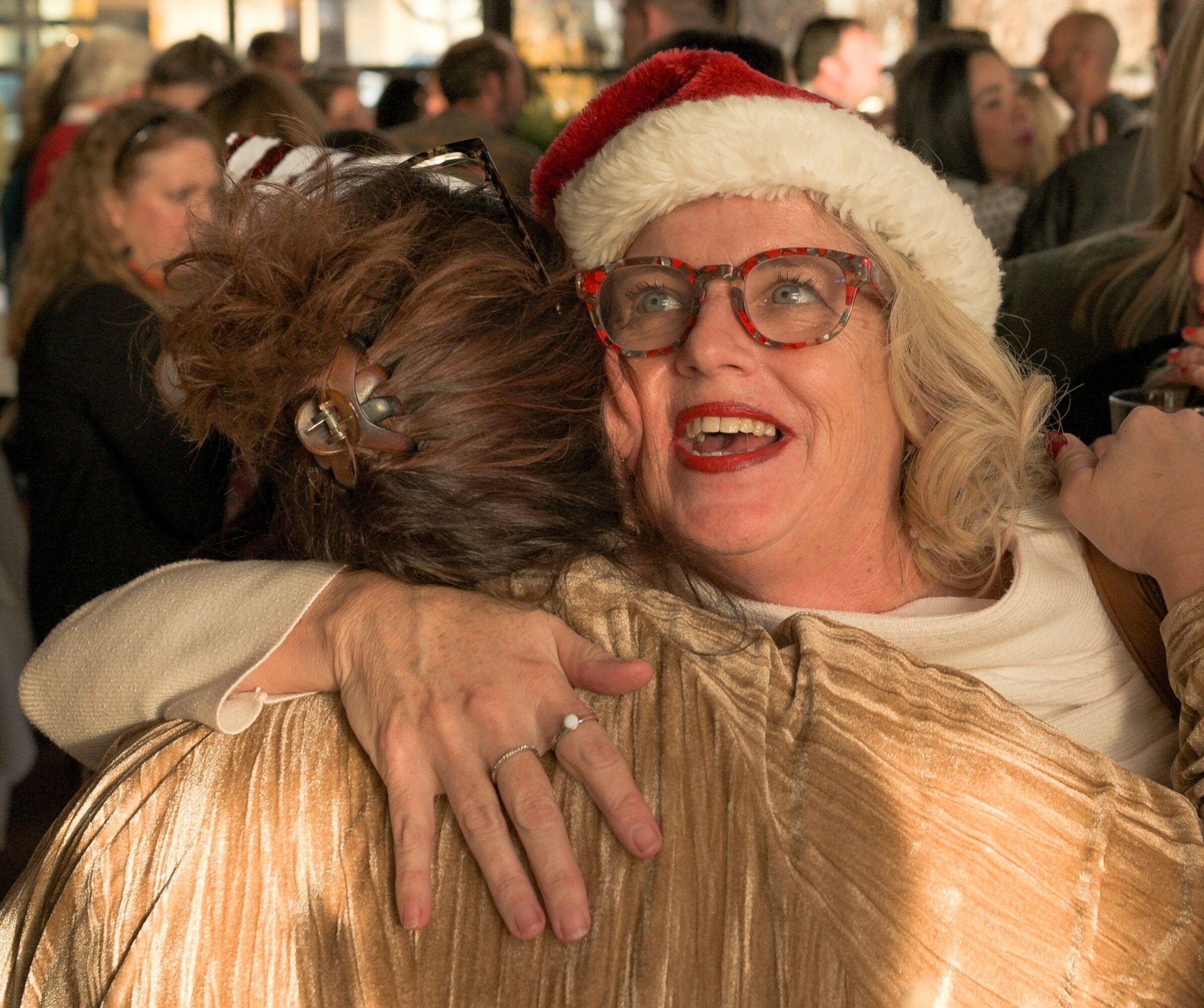 Two women hugging, one wearing a Santa hat and glasses, smiling warmly, surrounded by a crowd.