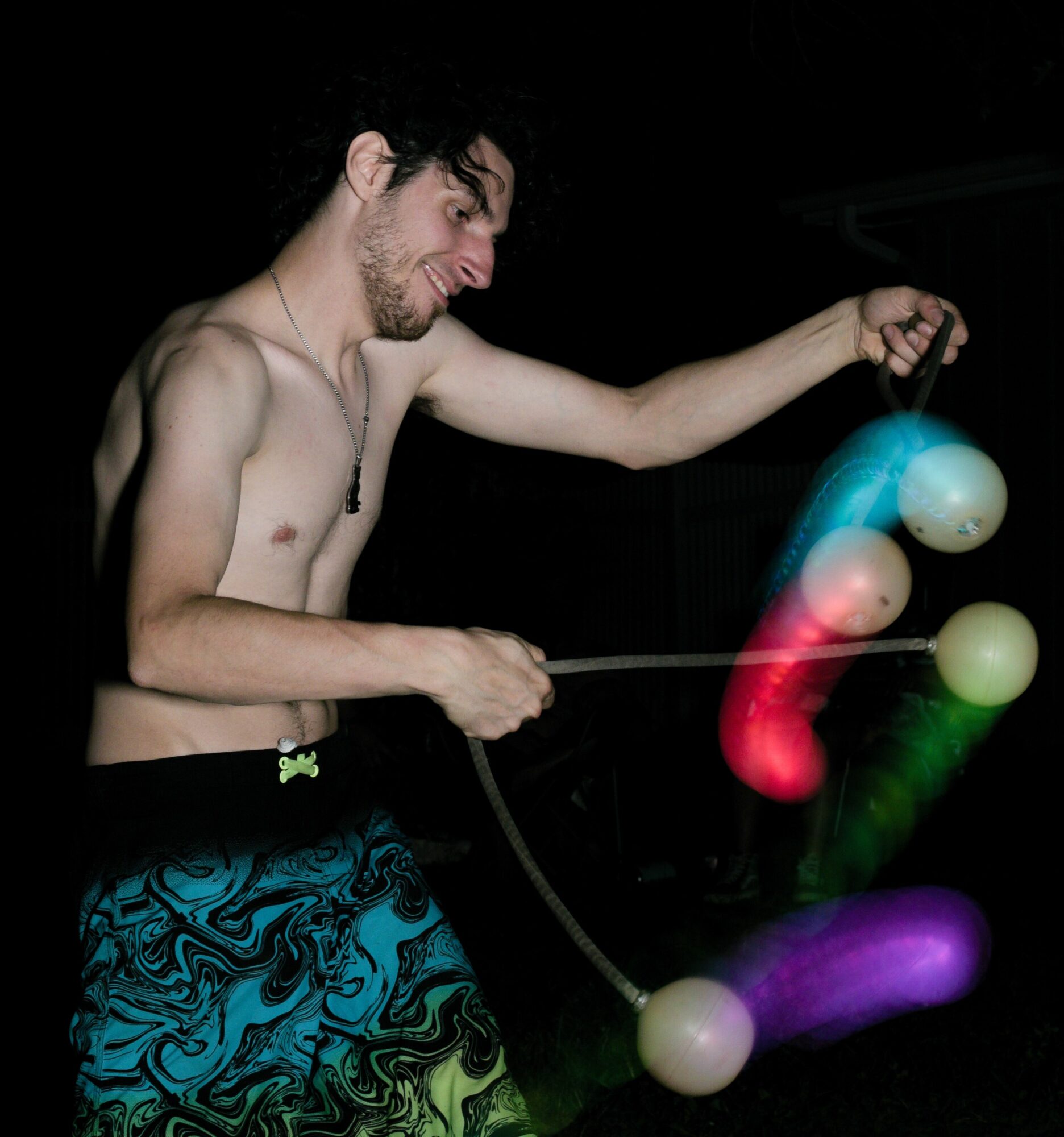 A shirtless man with glasses and dark hair juggles colorful balls in a dark setting.
