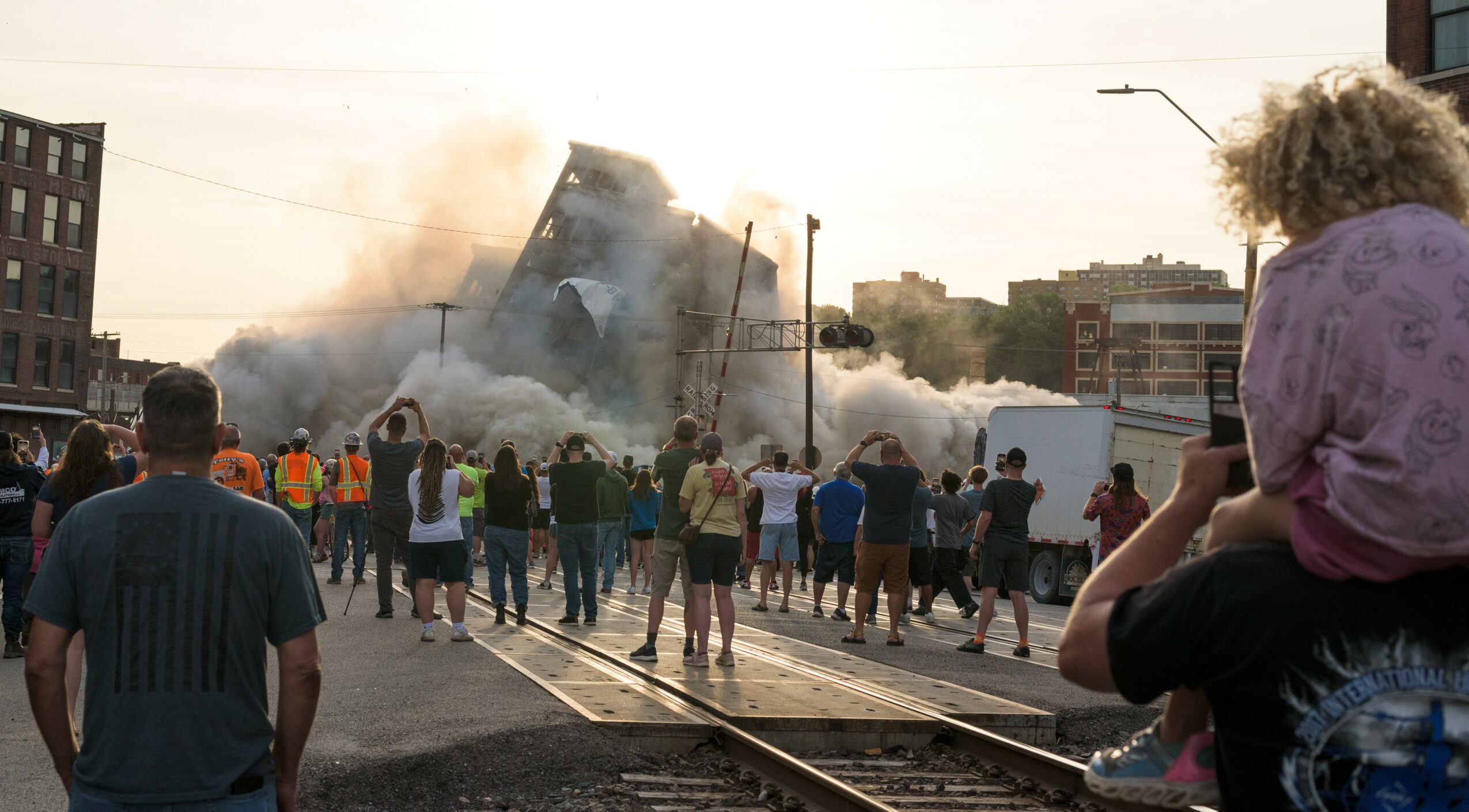 People gather on a street watching a building collapse with smoke and dust in the background.