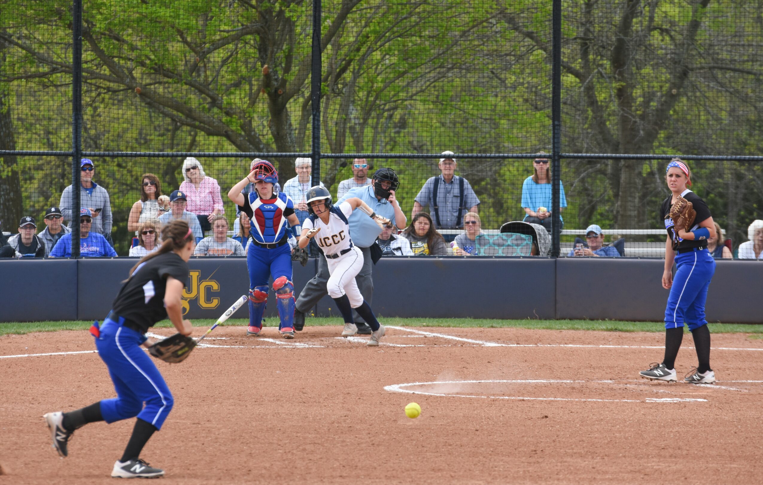 Softball game with players on field and spectators behind fence, yellow ball on ground, player preparing to bat.