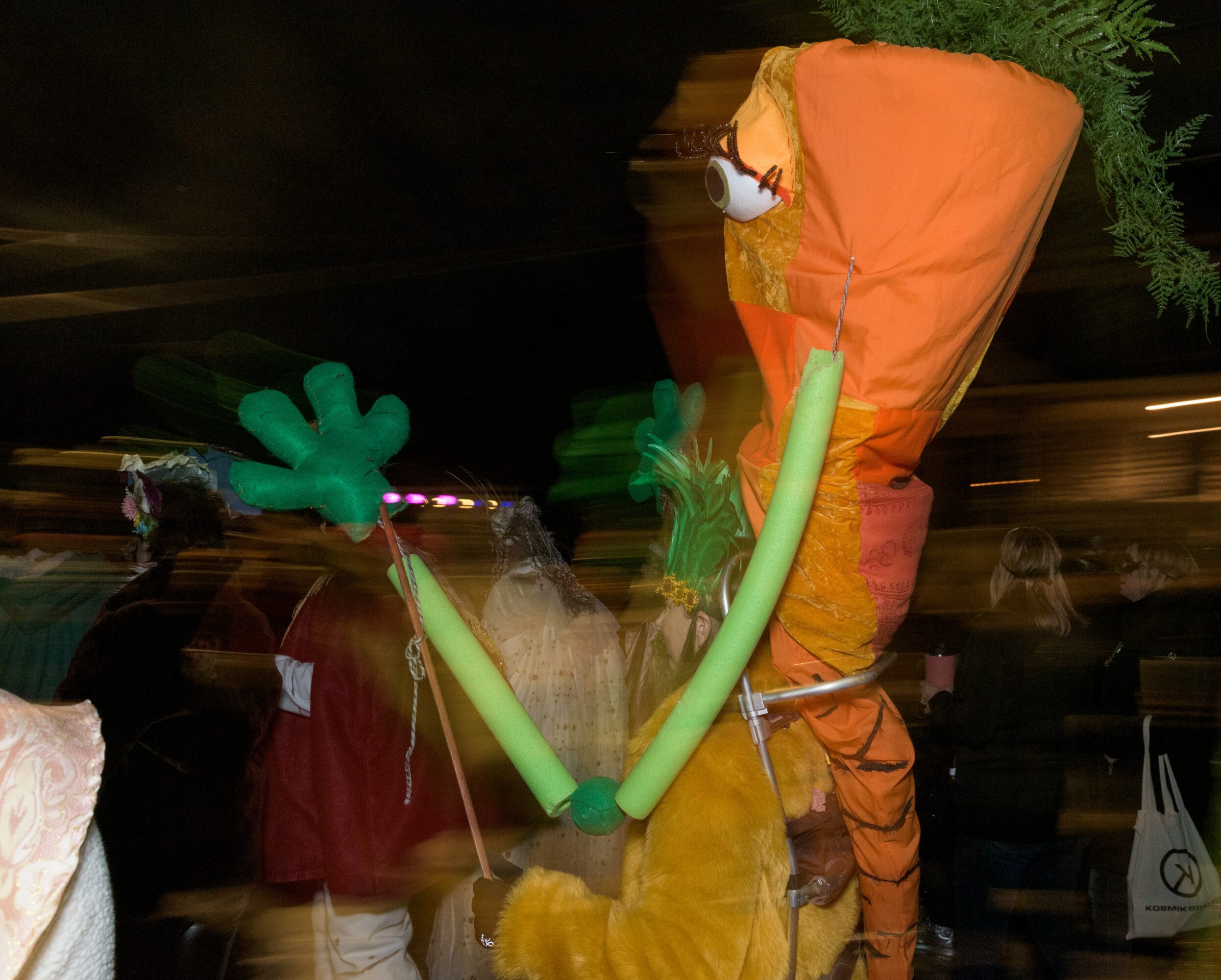 Person in a costume with large green gloves and an orange headpiece, moving in a crowded area at night.
