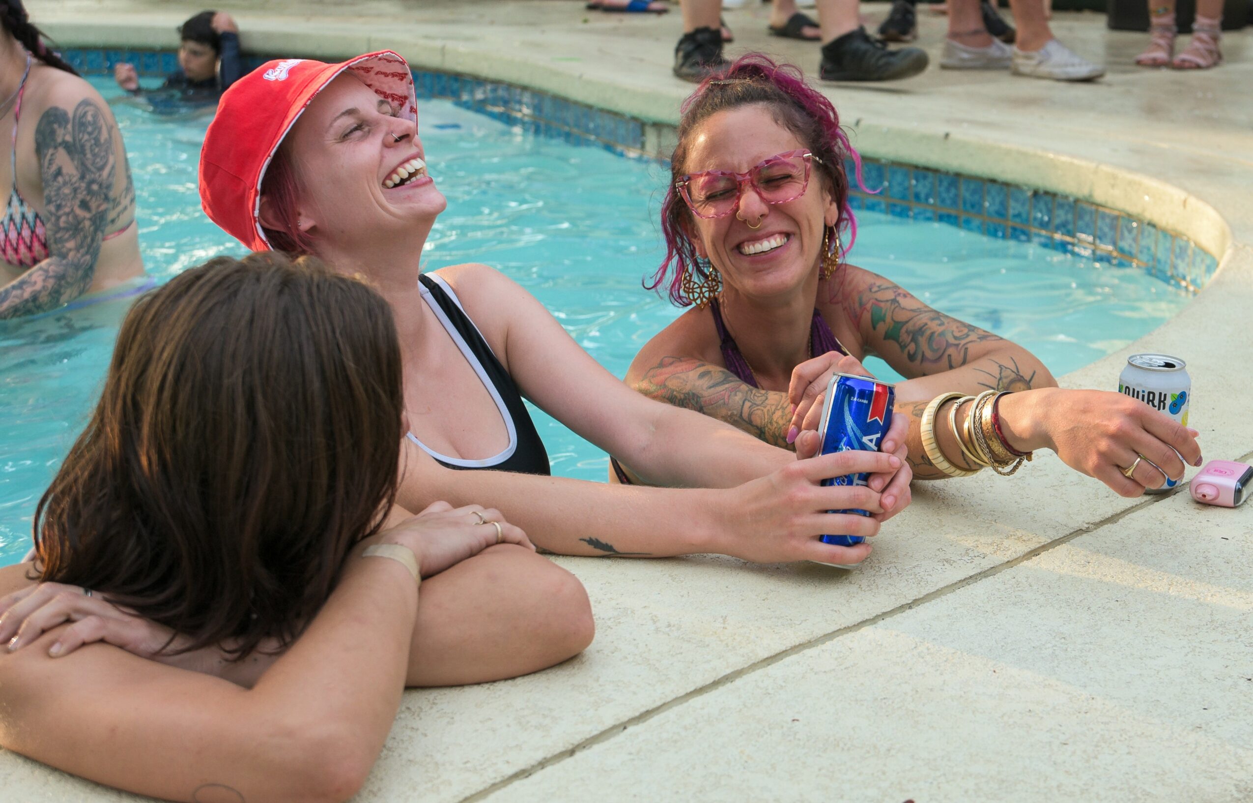 Three women smiling and laughing at the poolside, one holding a drink and a phone, with others in the background.