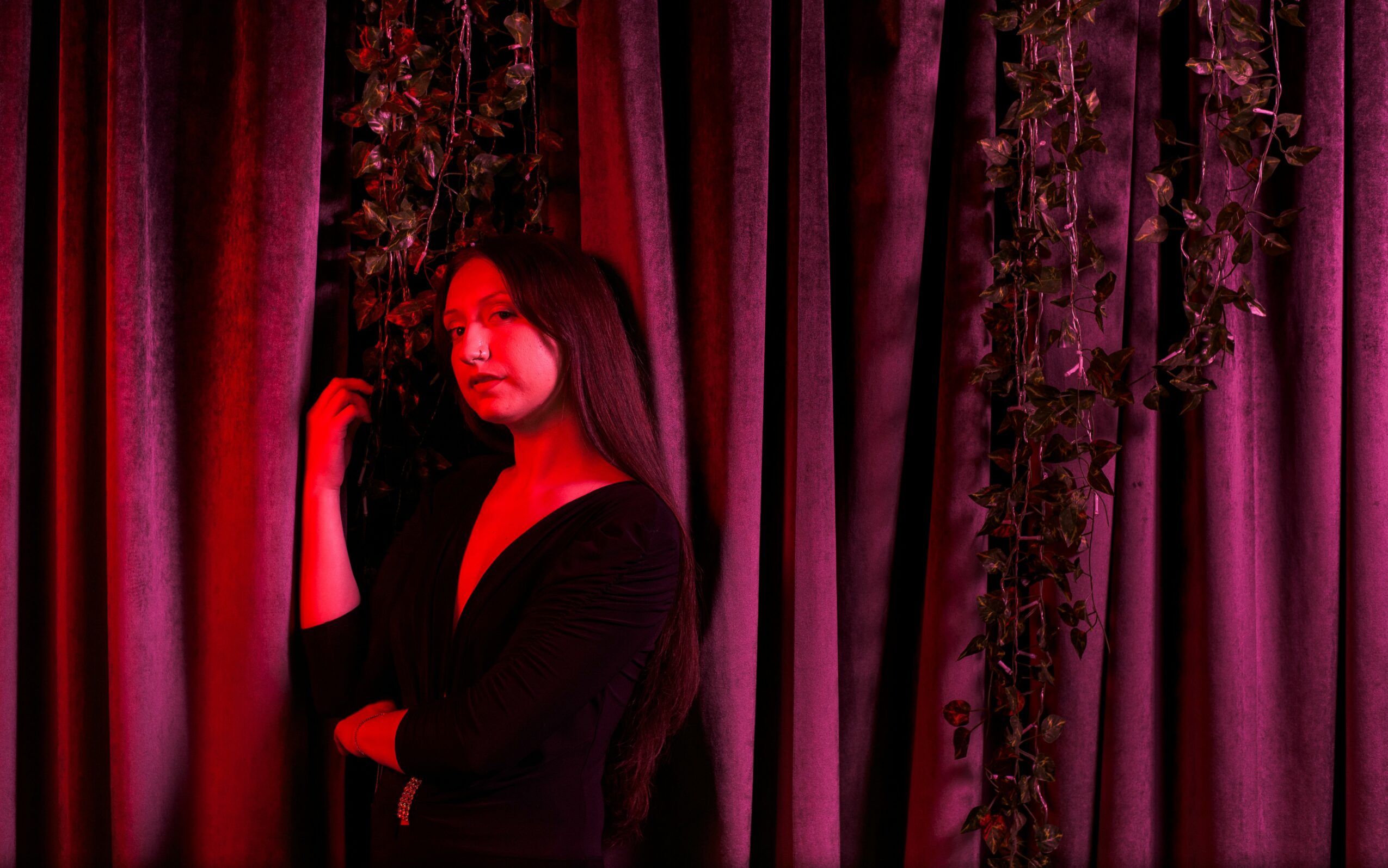 Woman with long dark hair standing in front of curtains with hanging vines, illuminated by red and purple lighting.