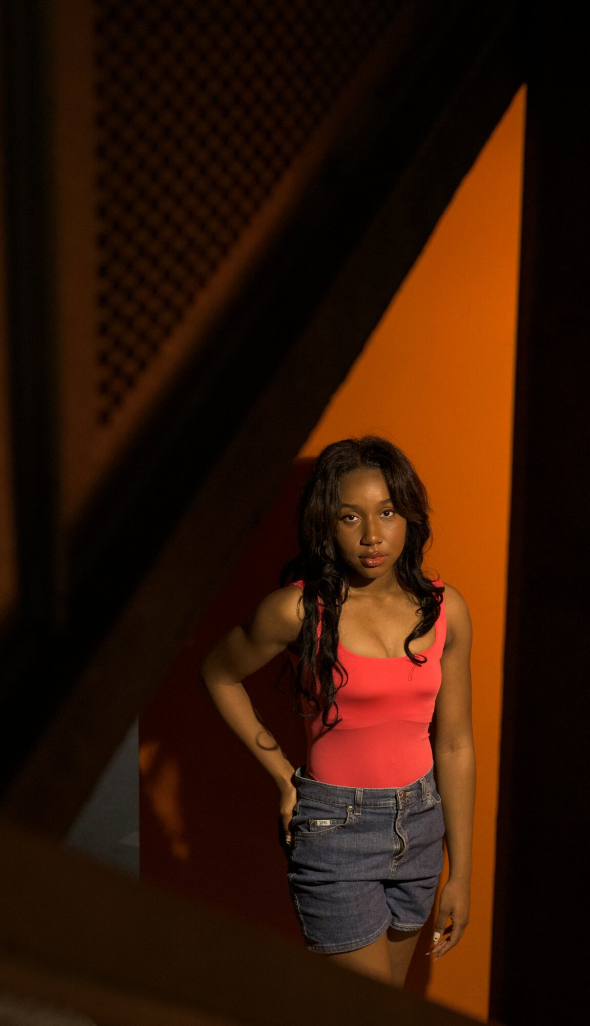 Young woman with long dark hair in a pink top and denim shorts stands against an orange background.
