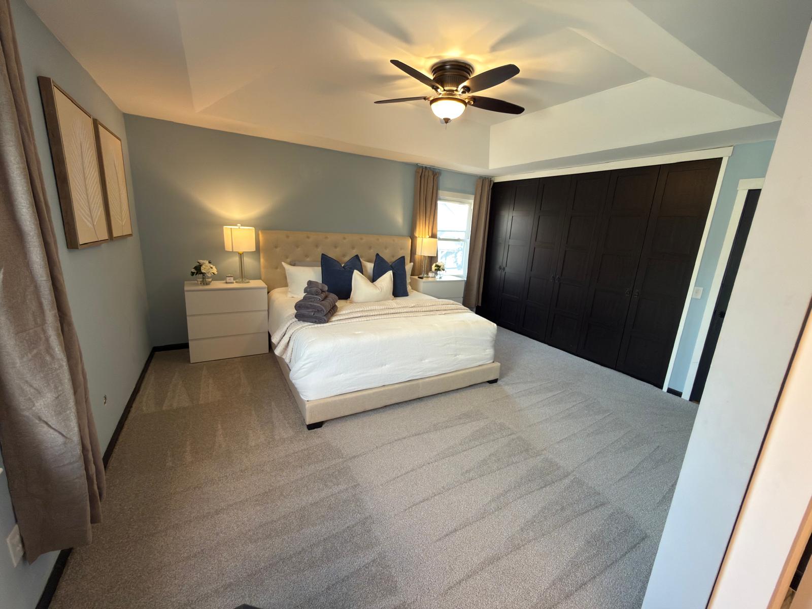 Bedroom with bed, nightstands, lamps, ceiling fan, and large dark closet doors, light-colored walls, and carpeted floor.