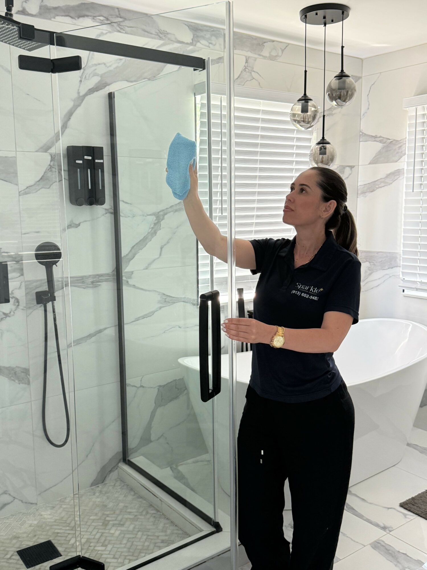 Woman cleaning glass shower door with a cloth in a modern bathroom with white marble walls and a bathtub.