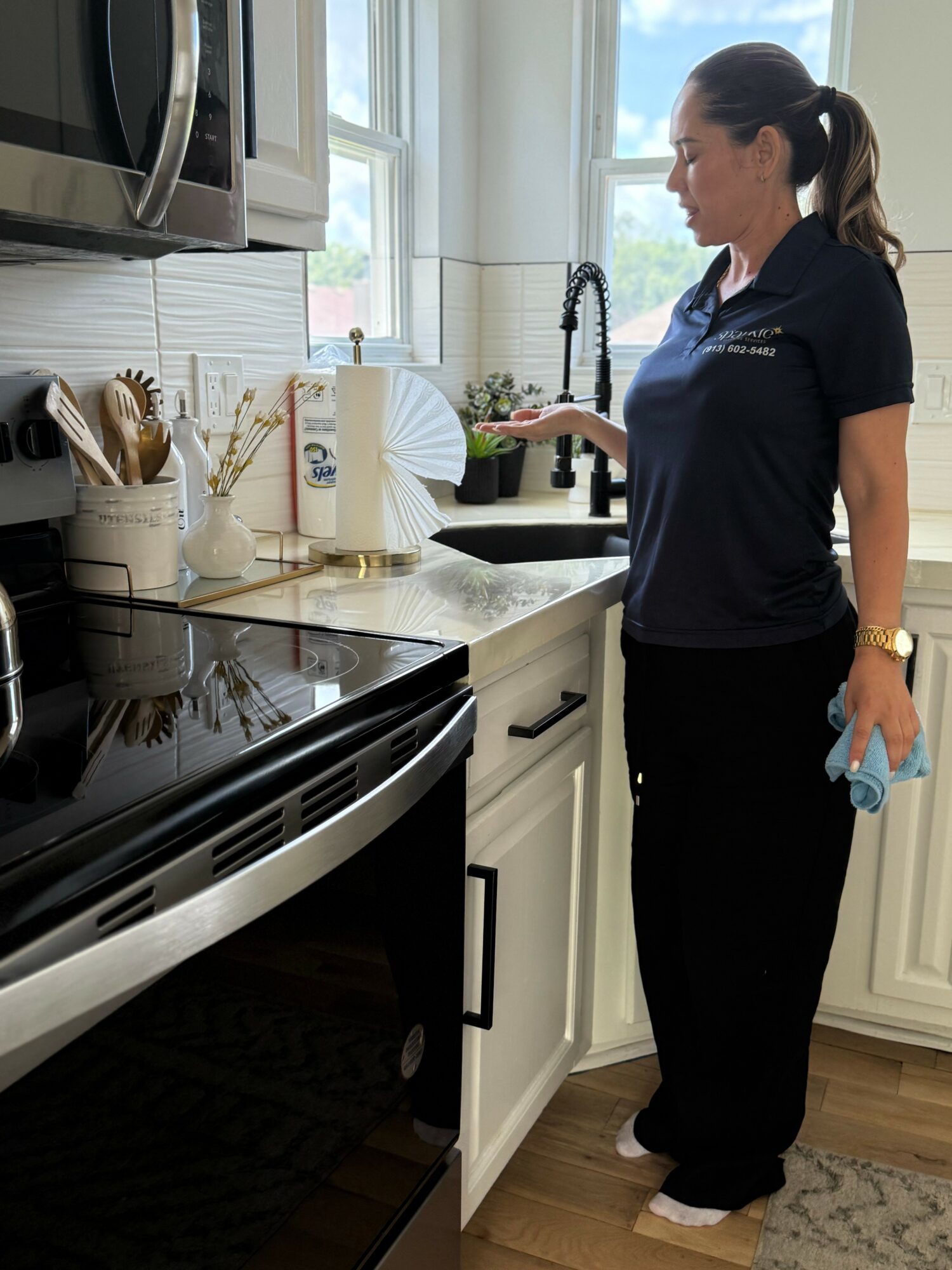 Woman in dark uniform cleaning kitchen counter near sink with window and appliances in background.