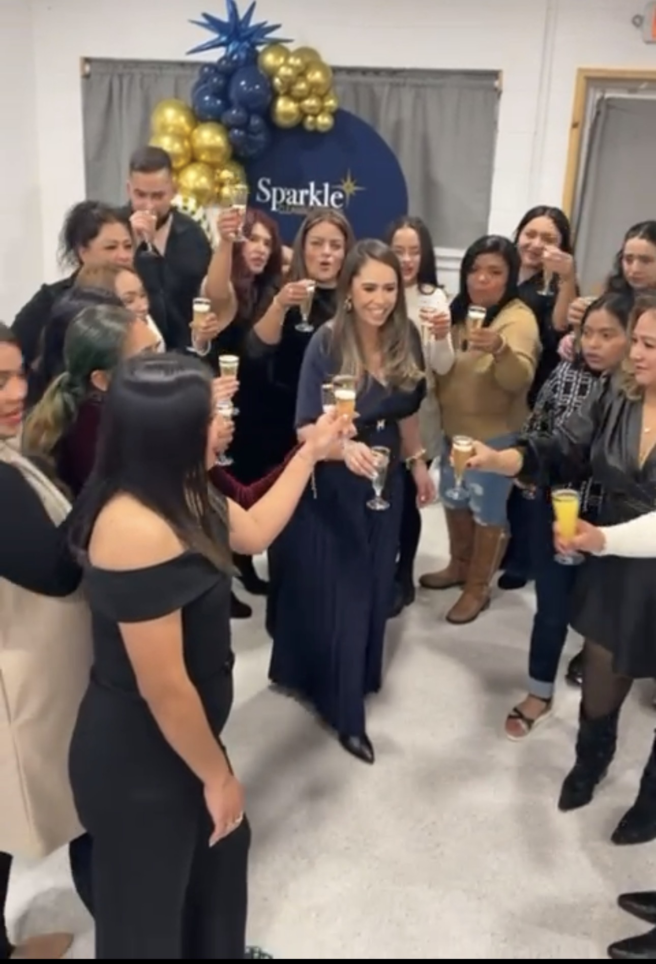 Group of people celebrating with drinks in front of a blue and gold balloon decoration and a sign that says 'Sparkle'.