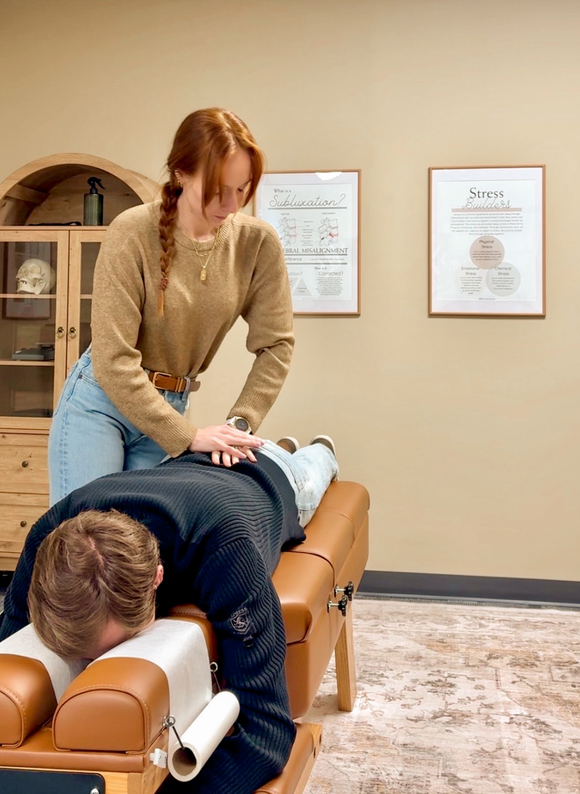 A woman performs a massage on a person lying face down on a massage table in a room with framed posters on the wall.