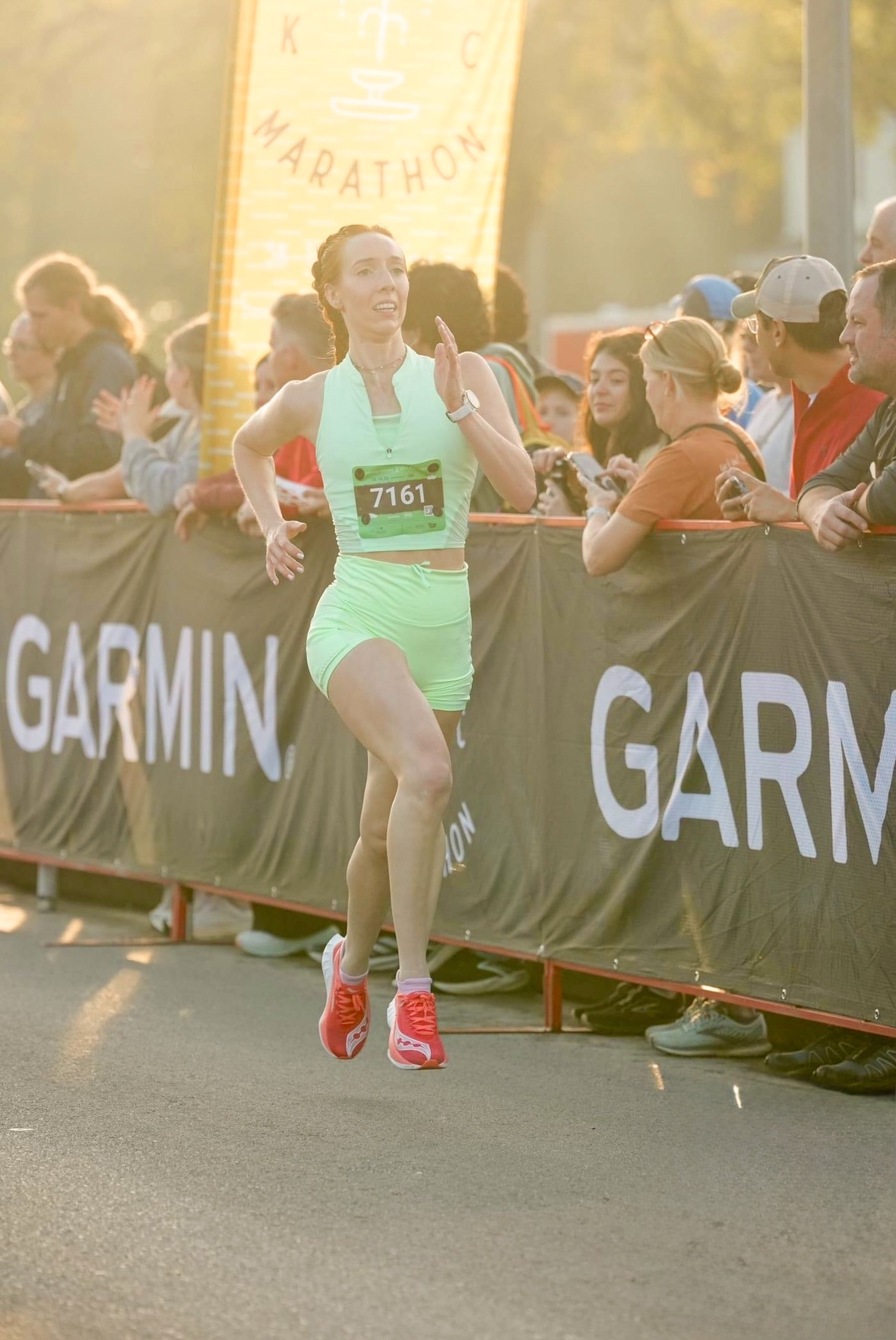 Female runner in green shorts and top crossing finish line with spectators on side, sunlight in background.