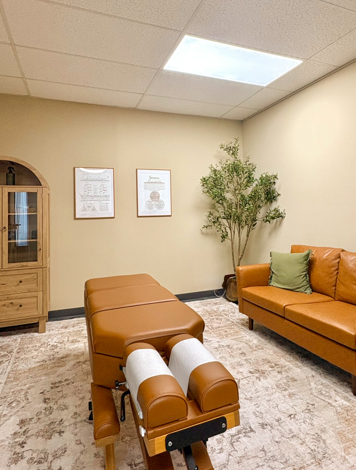 Room with beige walls, a skylight, a potted plant, a brown leather ottoman, and a matching sofa with a green pillow.