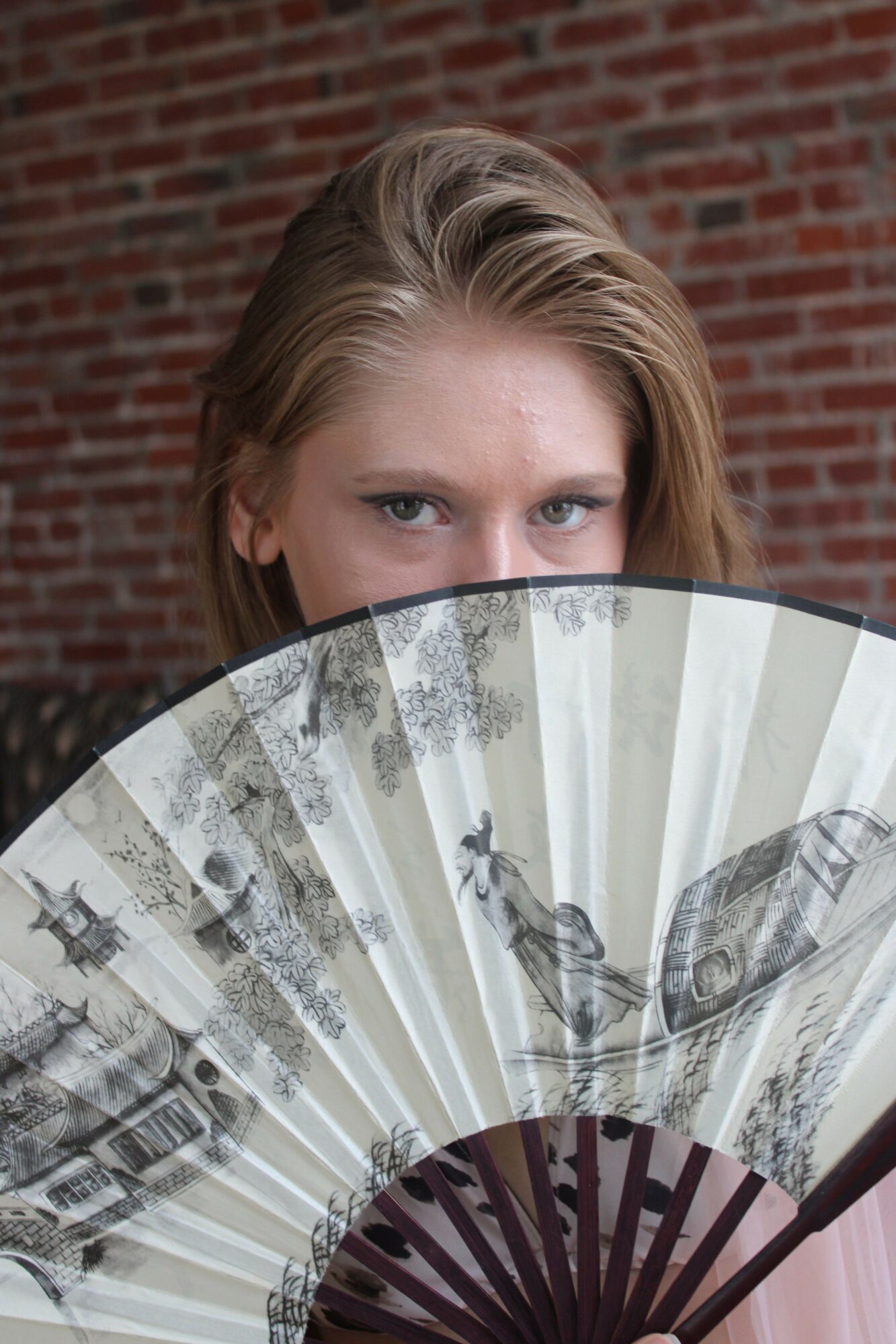 Partially visible woman with blonde hair and blue eyes holding a decorative fan, brick wall background.