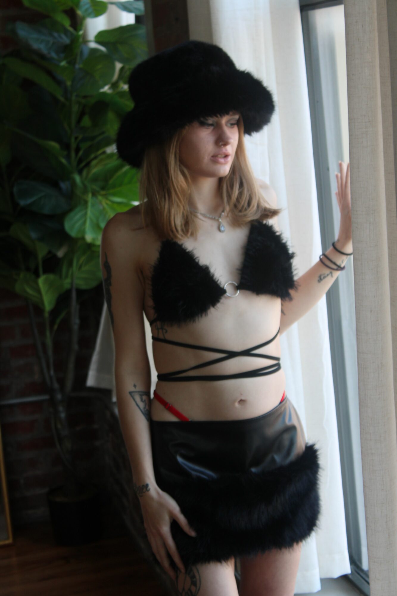 Person in black furry outfit with straps, standing indoors near large green plants, with a window in the background.