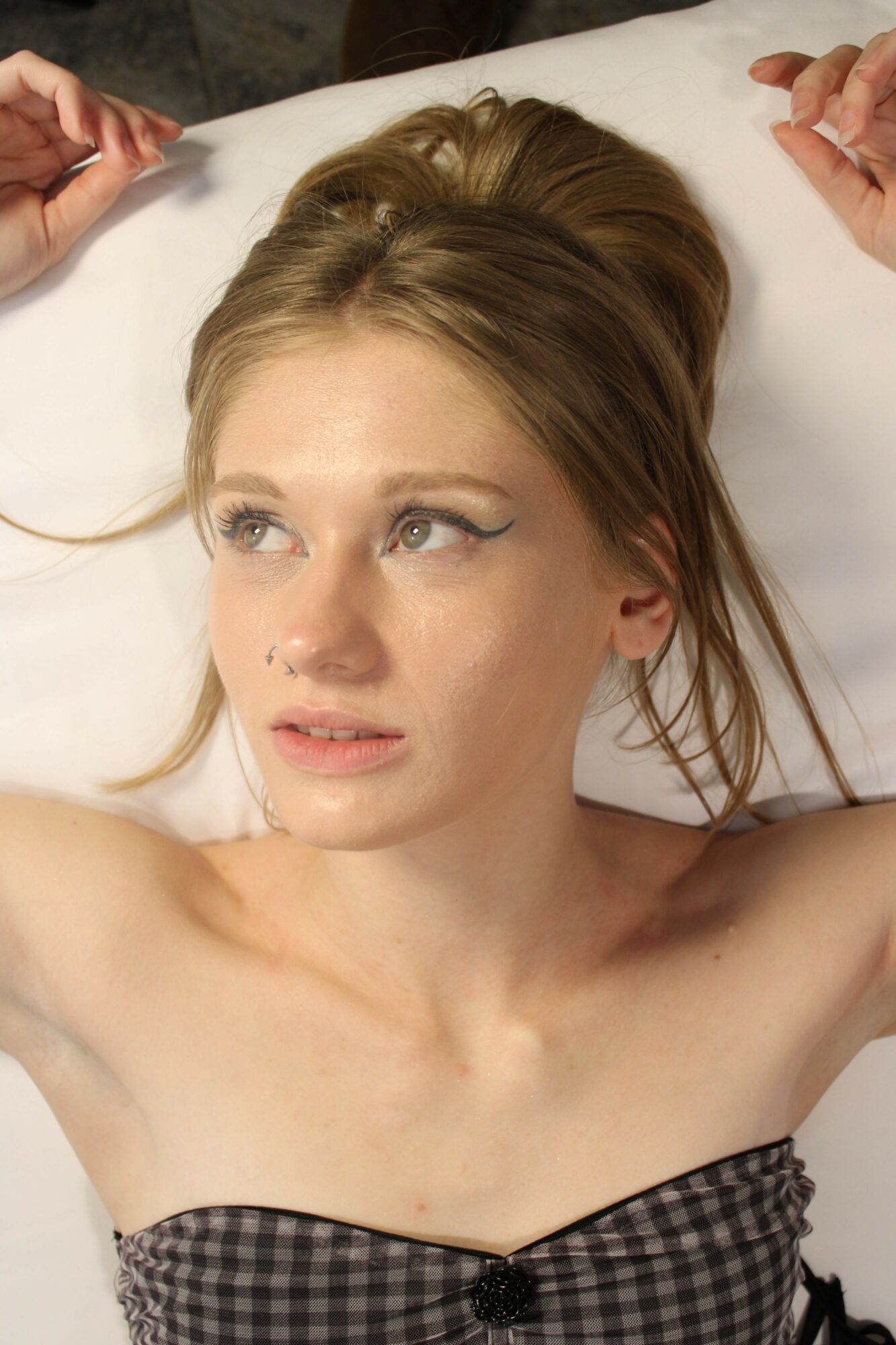 A woman with light brown hair lying on a white surface, wearing a checkered top, looking to the side.