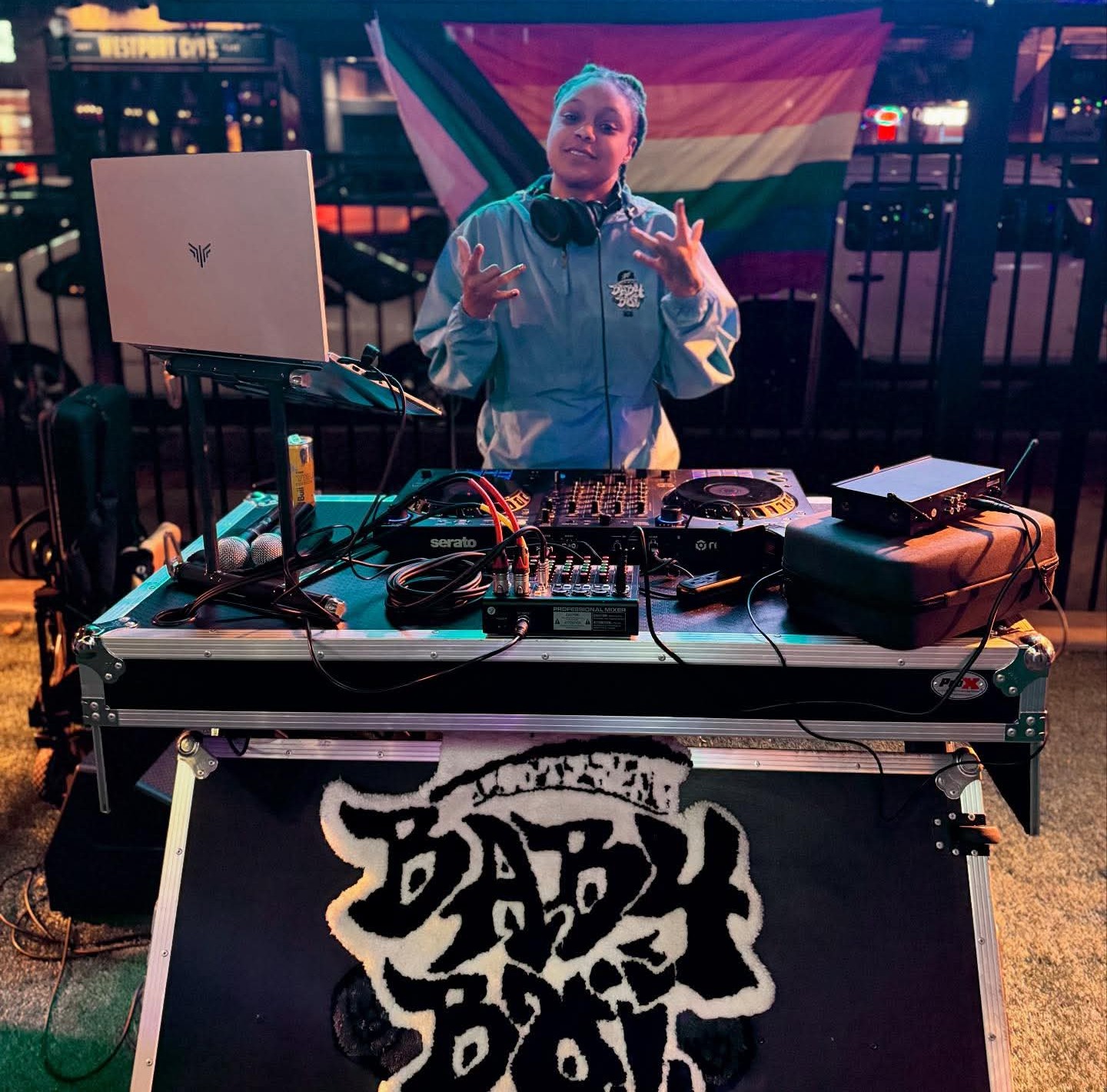 Person standing behind DJ equipment with a rainbow flag in the background, making hand gestures, in a dimly lit setting.