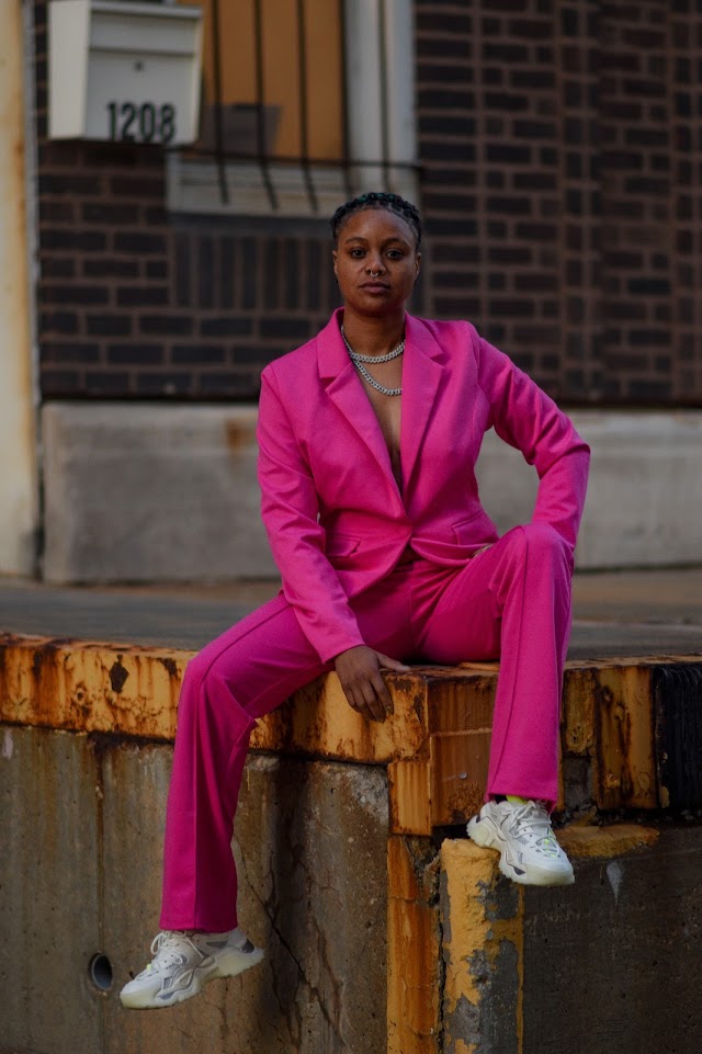 Woman in pink suit sitting on a ledge outdoors, brick building background, white sneakers, confident pose.