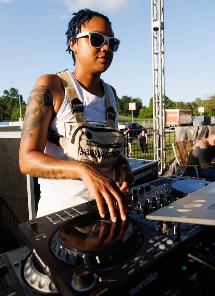 Person with sunglasses operating DJ equipment outdoors, wearing a white tank top and tattoos on arm.