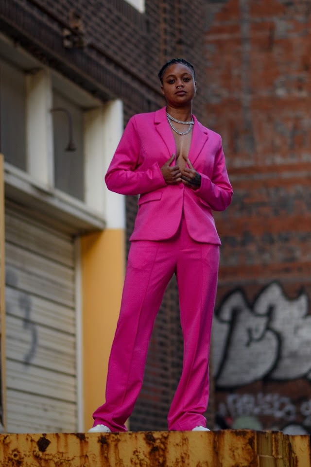 Woman in a bright pink suit standing outdoors in an urban setting, with graffiti and brick walls behind her.