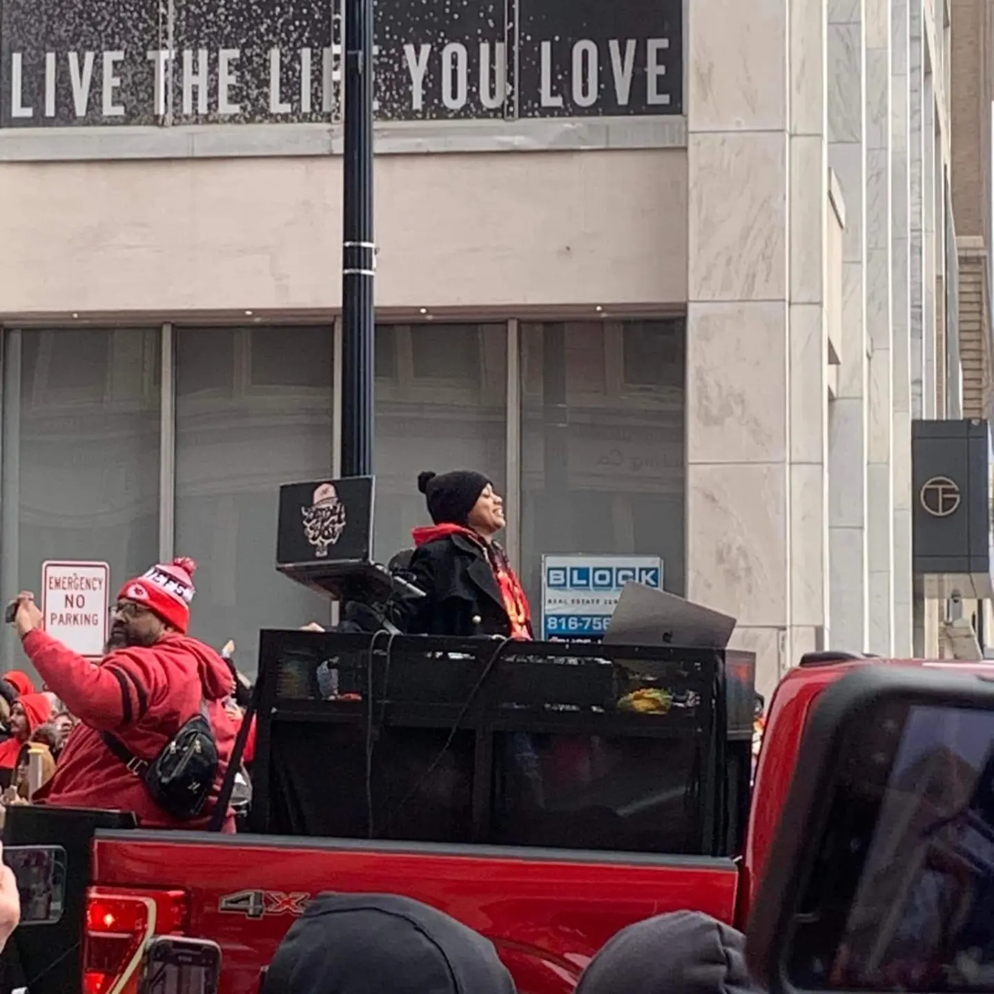 Person standing on a vehicle with a black jacket and red scarf, surrounded by people in red clothing, in an urban setting.
