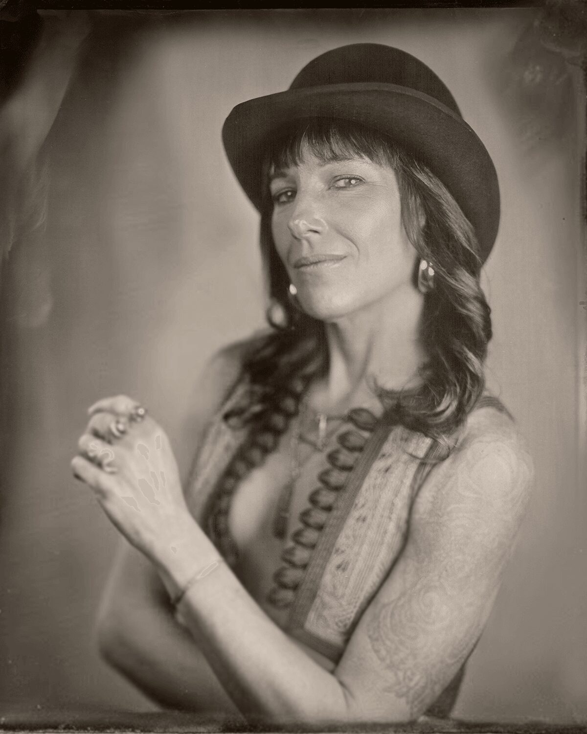 Woman wearing a hat, with long hair, jewelry, and a confident expression, posed with crossed arms.