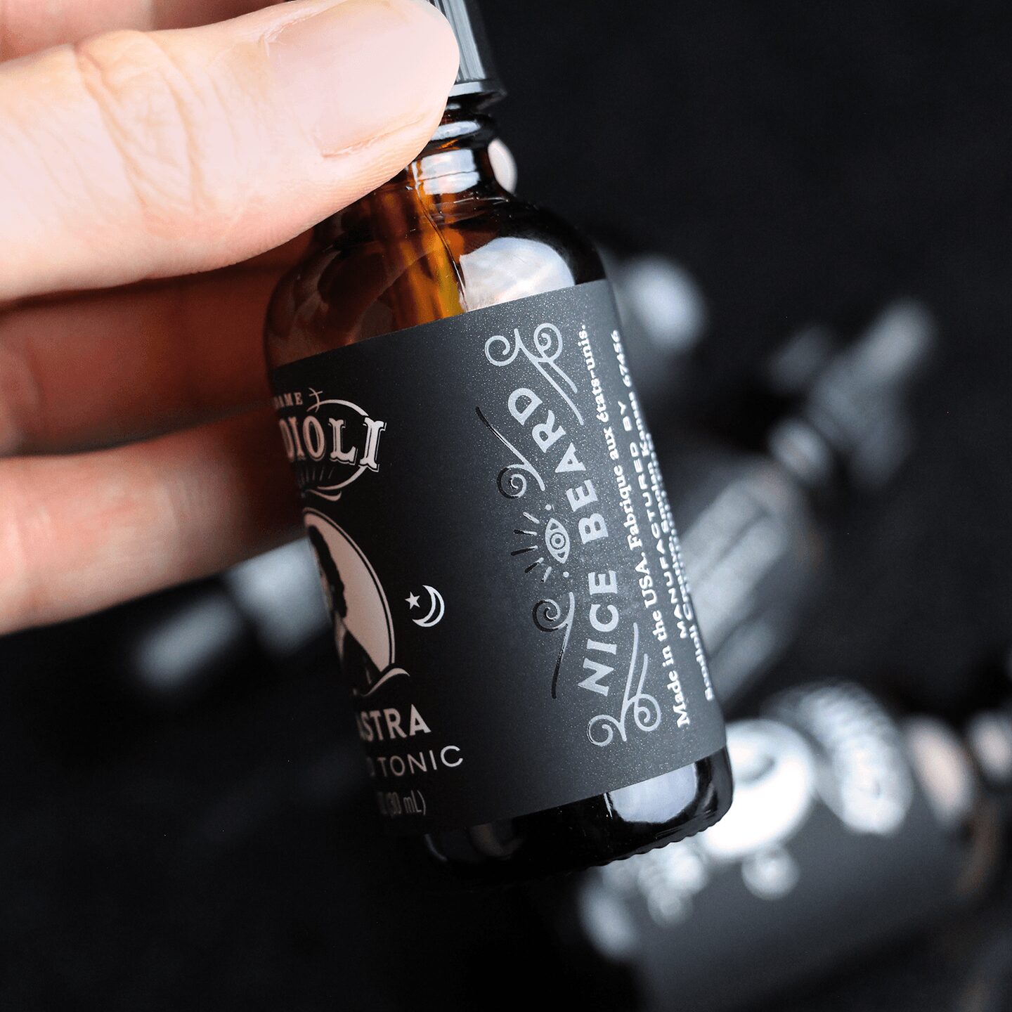 Hand holding a small amber glass bottle with a black label and dropper, with other bottles in the background.