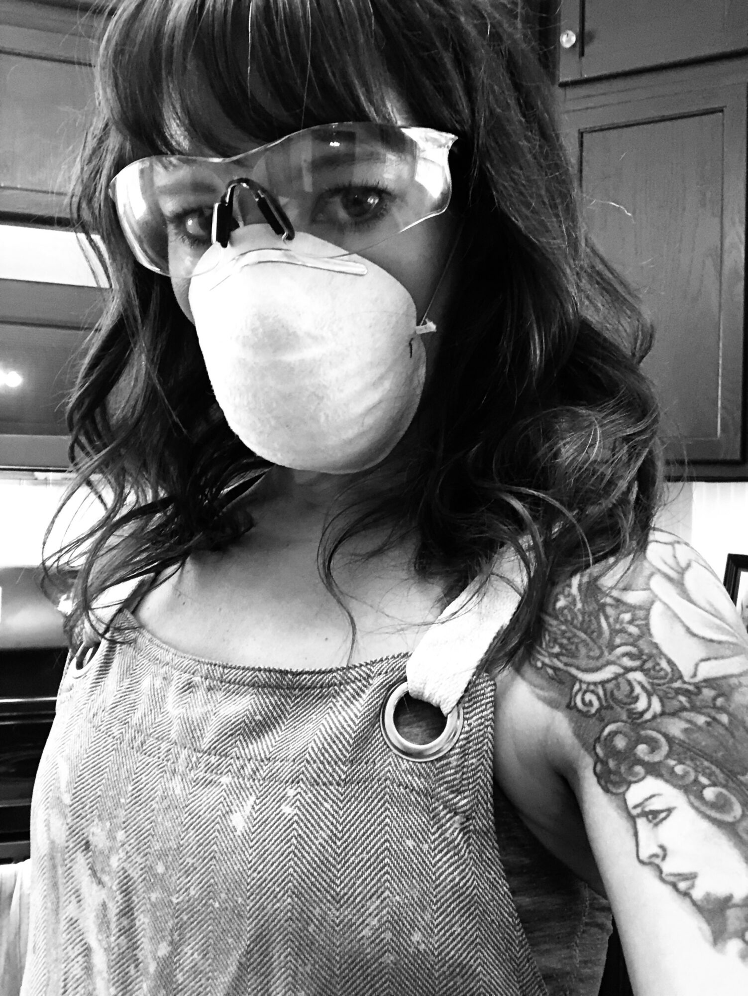 Woman with glasses and face mask, curly hair, tattoo on arm, wearing sleeveless top, in indoor setting.