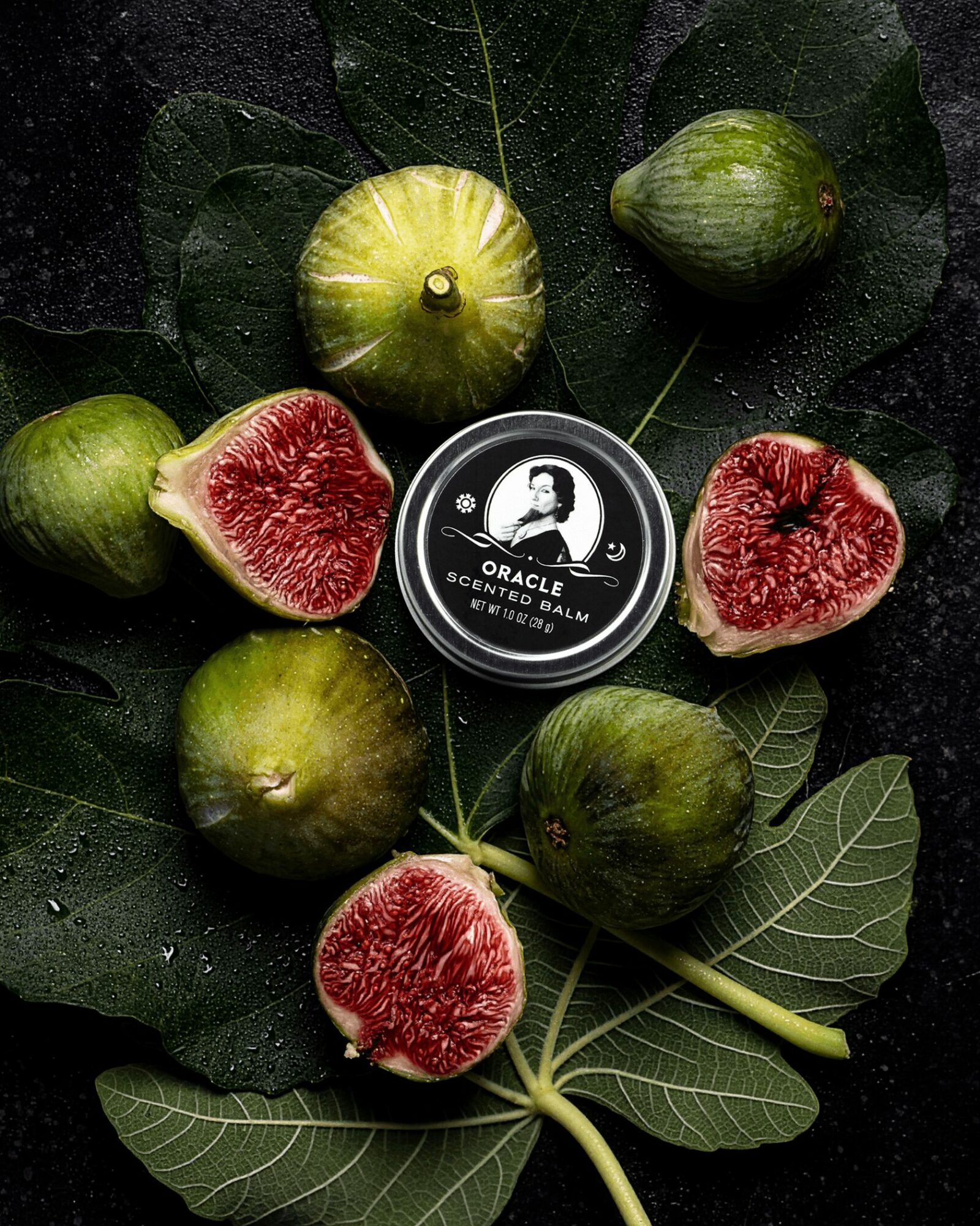 Figs and leaves arranged around a small container with a black and white logo, on a dark surface.