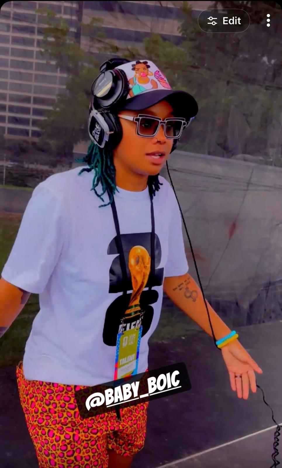 Person with sunglasses, colorful hat, and headphones, wearing a white shirt and patterned pants, standing outdoors.