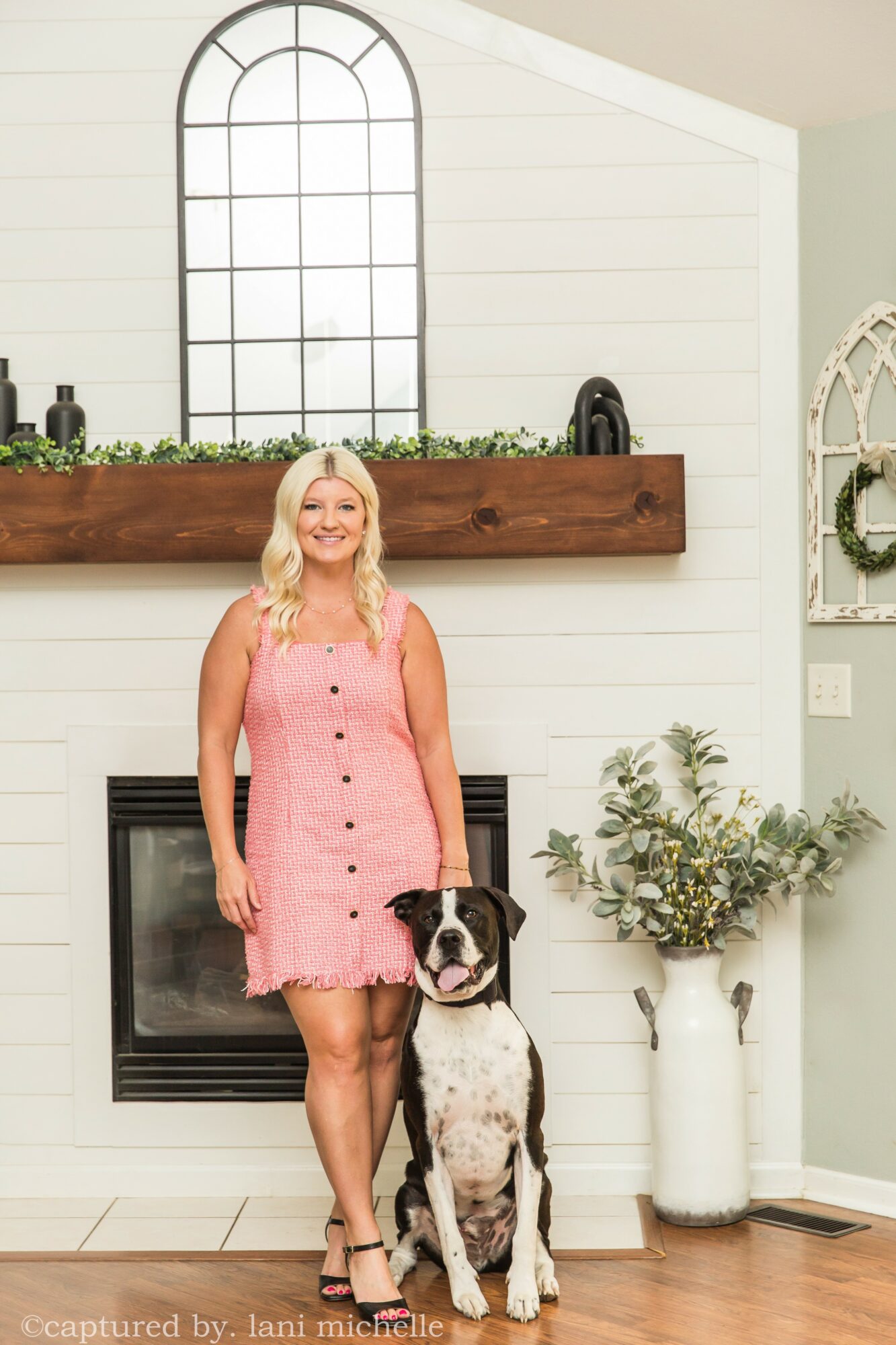 Woman in pink dress standing next to a large dog in a living room with a fireplace and decorative plants.