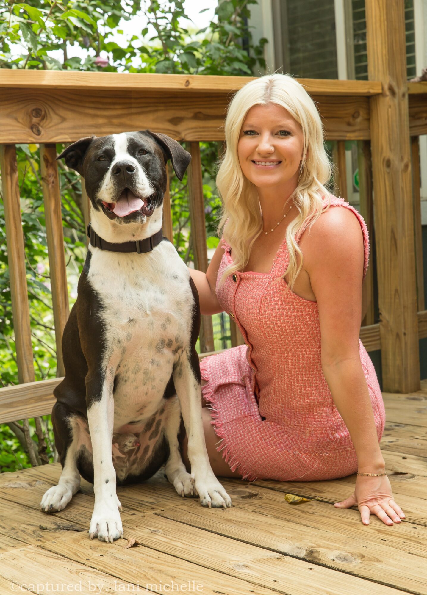 Woman with long blonde hair sitting on wooden deck next to a large black and white dog, smiling outdoors.