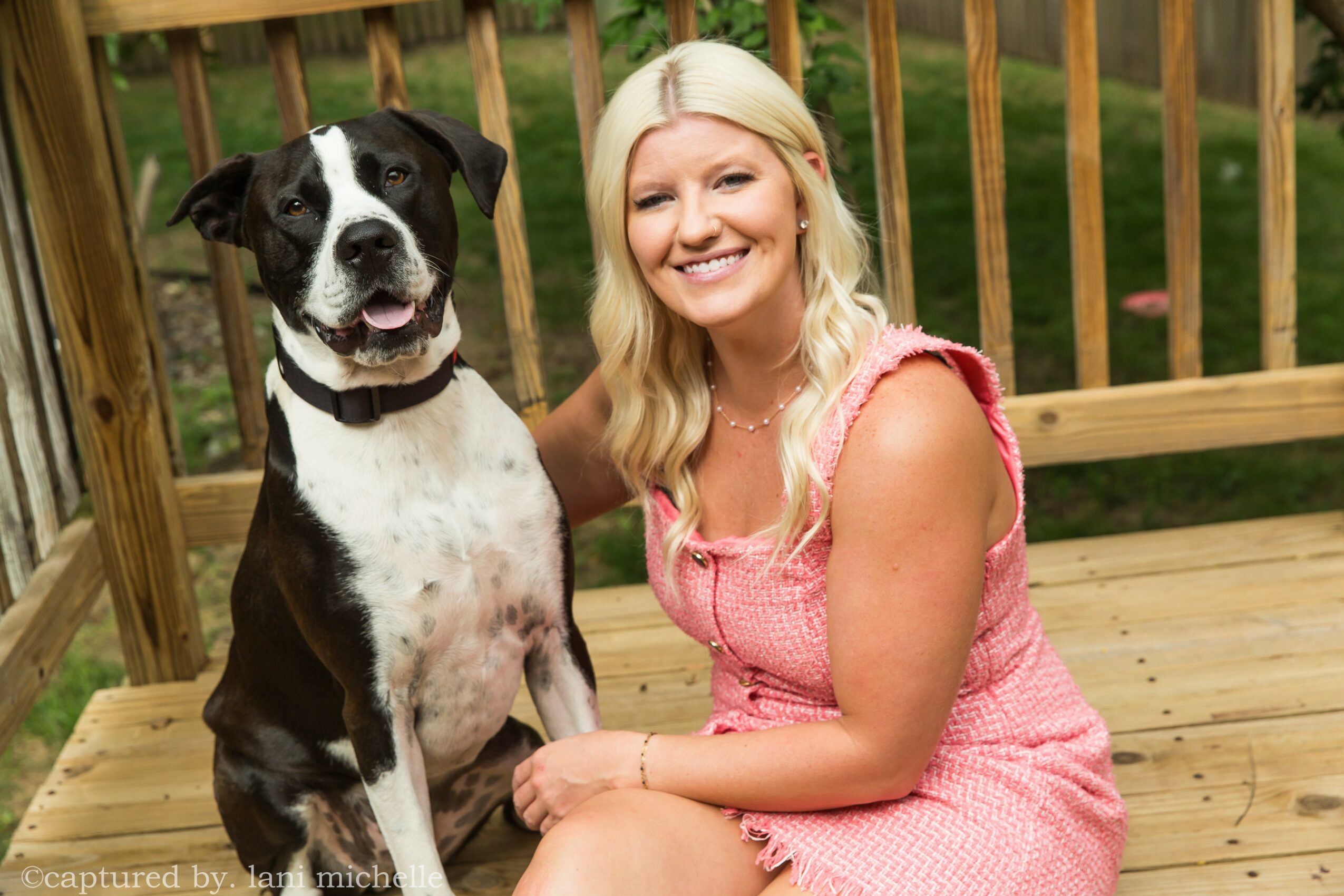 Woman with blonde hair in pink dress sitting on wooden deck next to a black and white dog, smiling outdoors.