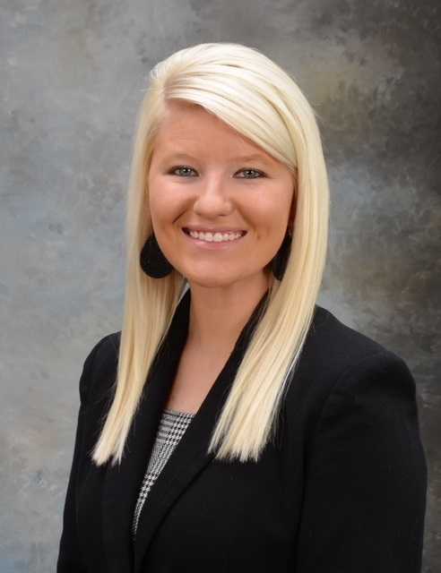 Portrait of a smiling woman with long blonde hair wearing a black blazer and patterned top, against a neutral background.