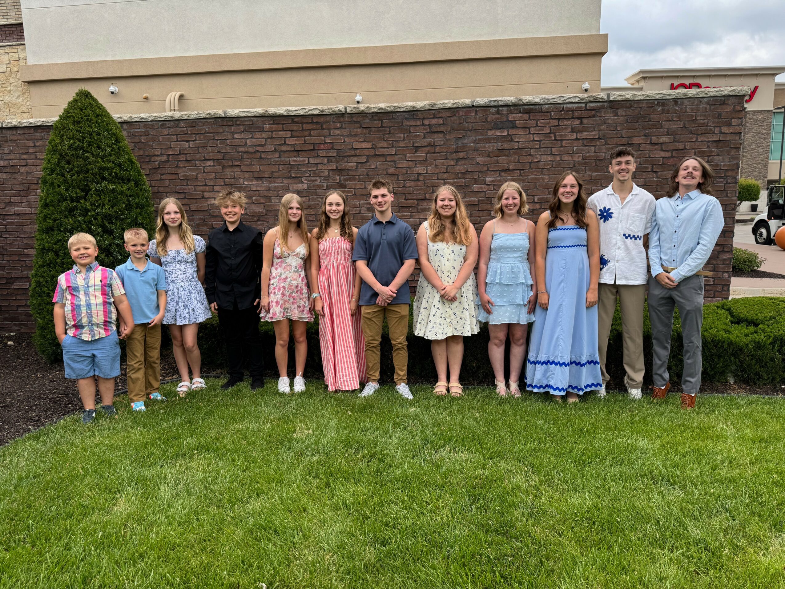 Group of children and teenagers standing on grass in front of a brick wall, dressed in casual and semi-formal clothing.