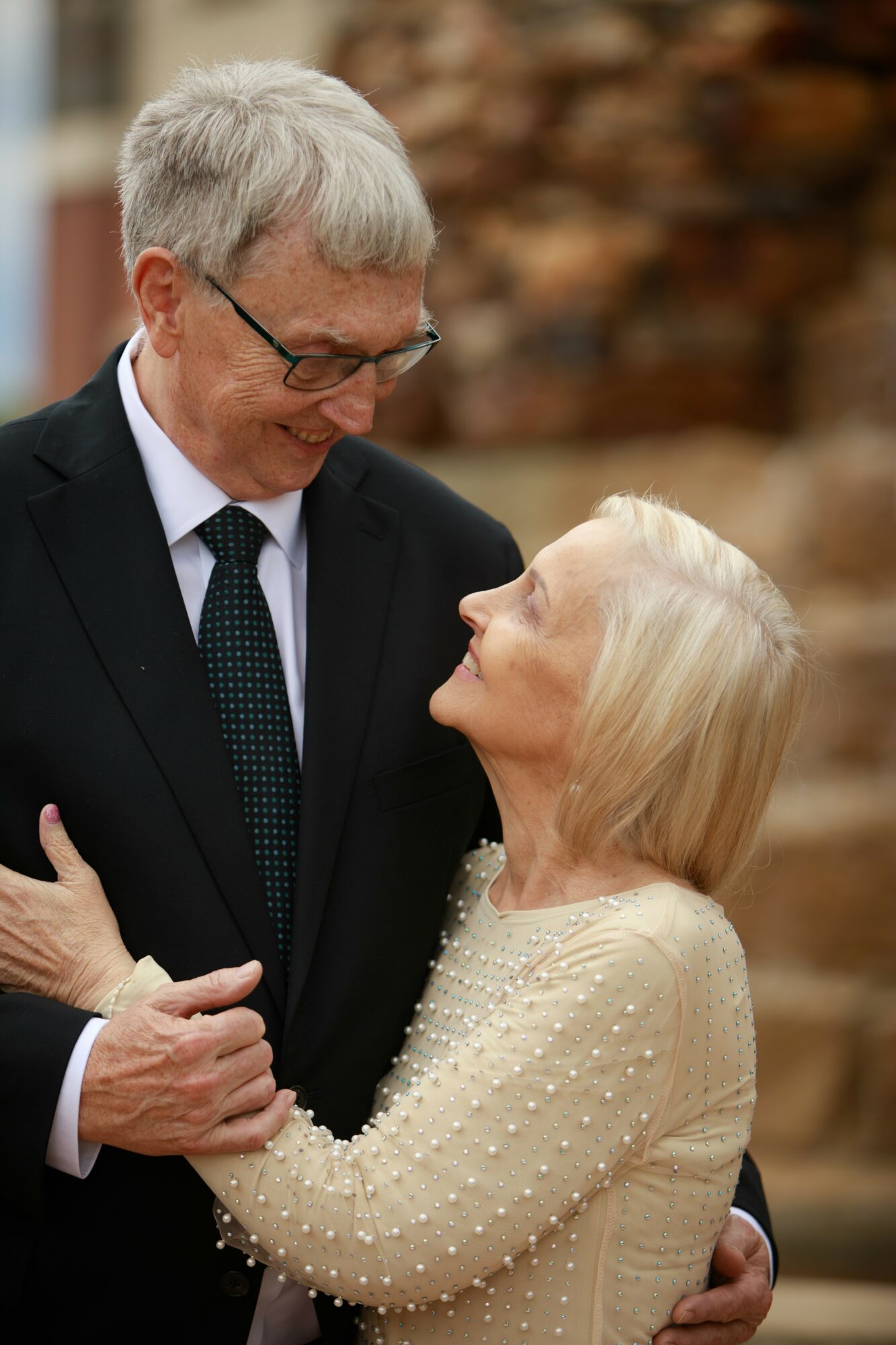 An elderly man and woman look at each other affectionately outdoors, smiling, with blurred autumn leaves in background.