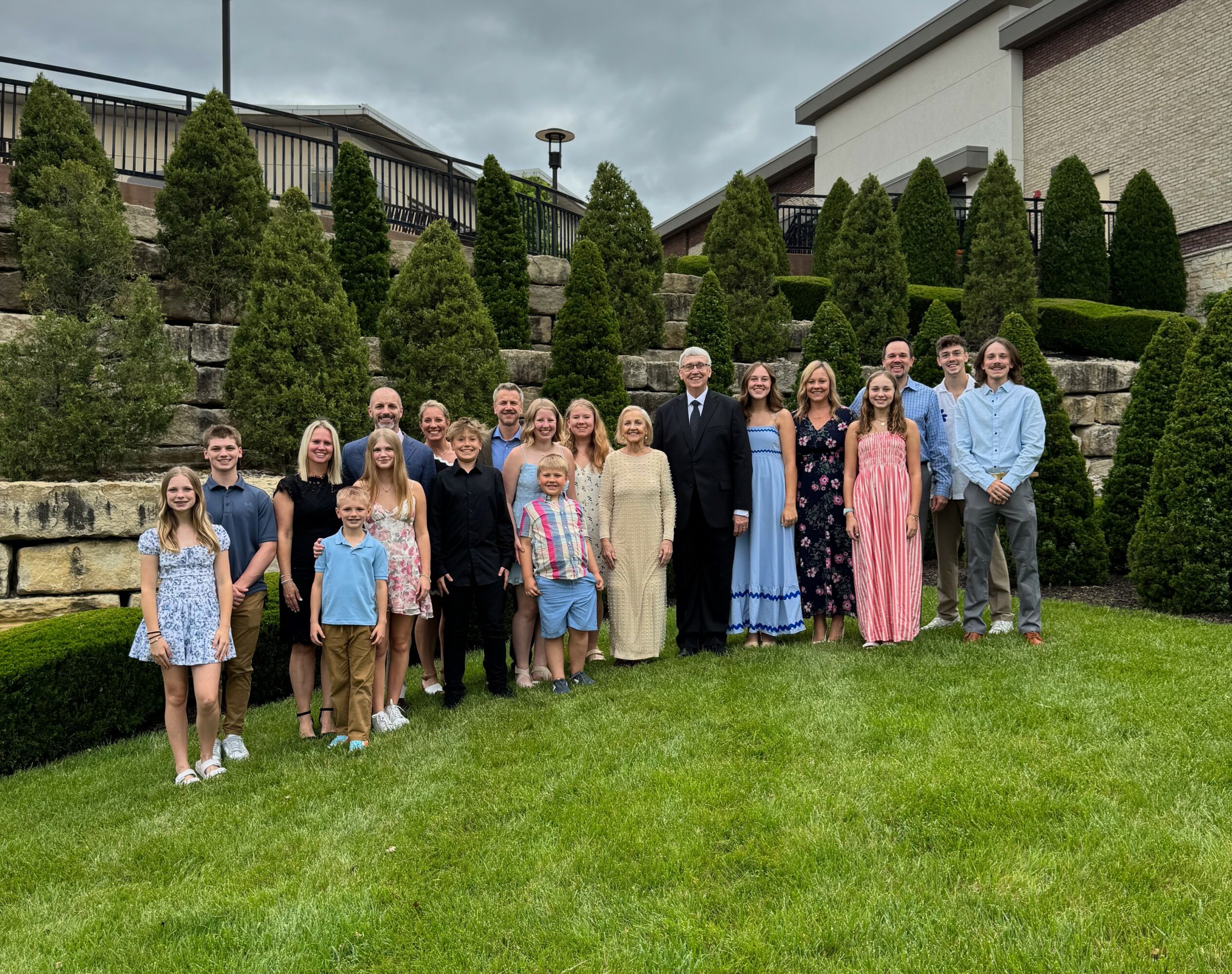 Group of people standing on grass in front of trimmed bushes and buildings, posing for a photo outdoors.
