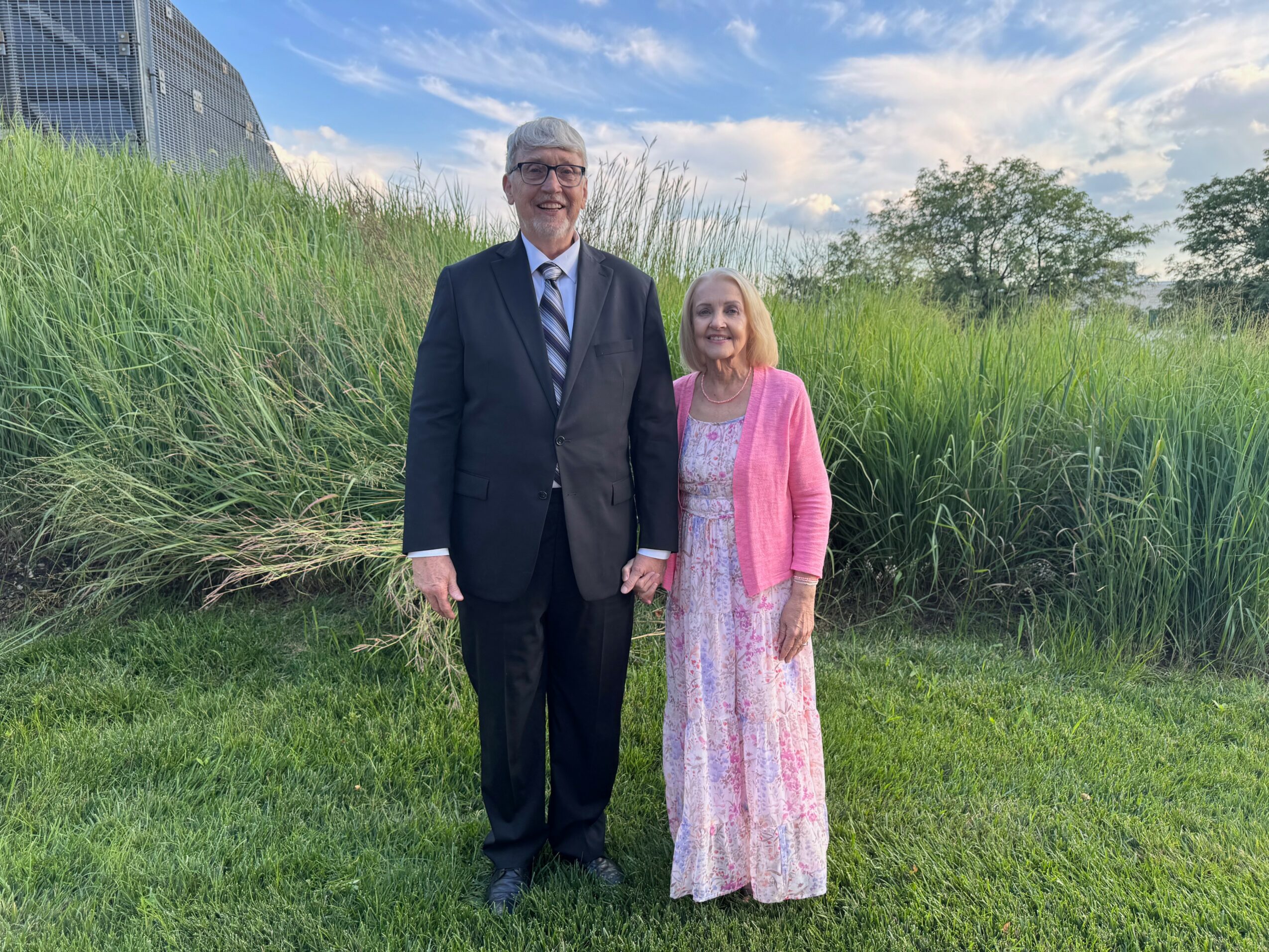 A man and woman stand outdoors on grass with tall plants and trees behind them, smiling at the camera.