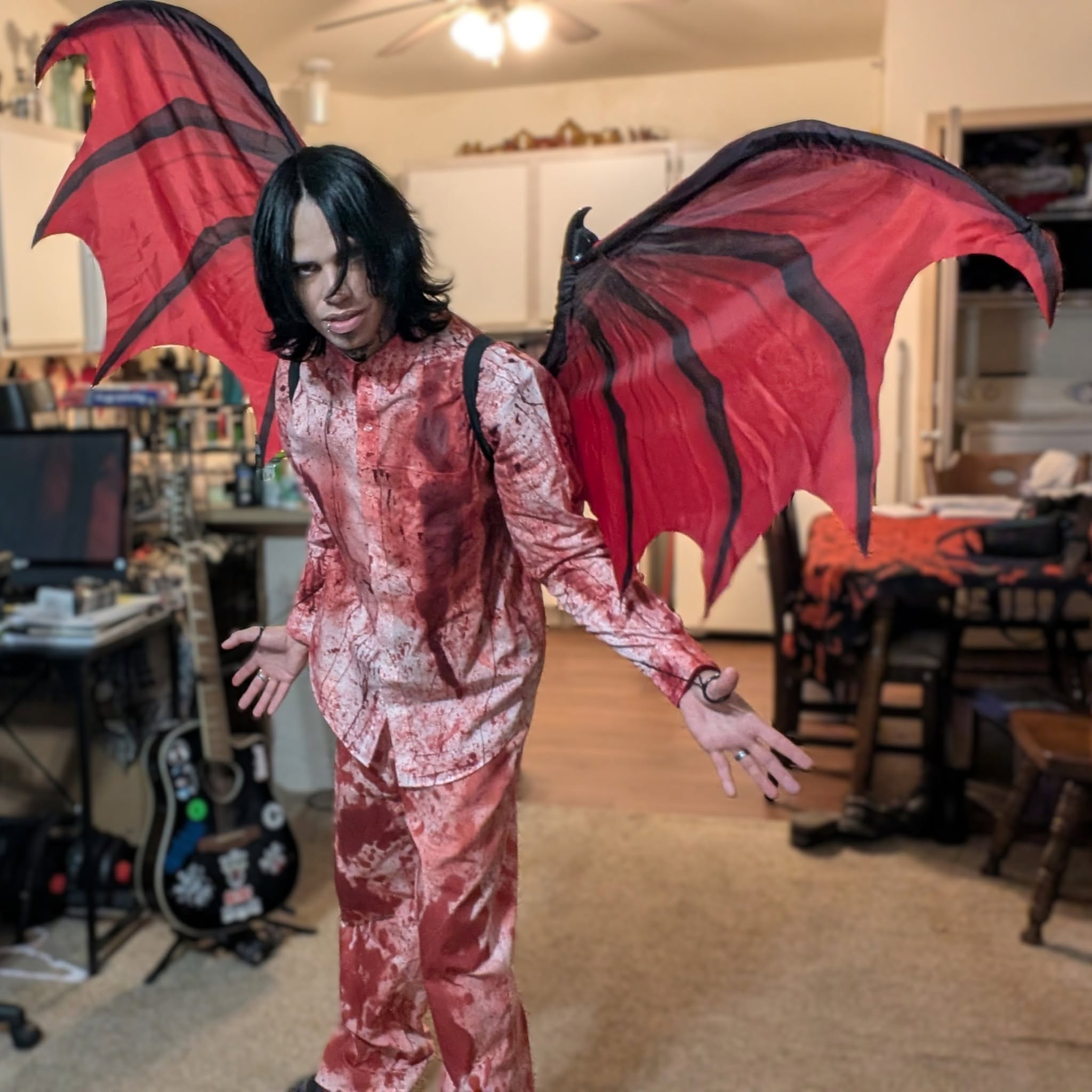 Person with red and black bat-like wings costume, standing indoors in a room with furniture and electronics.